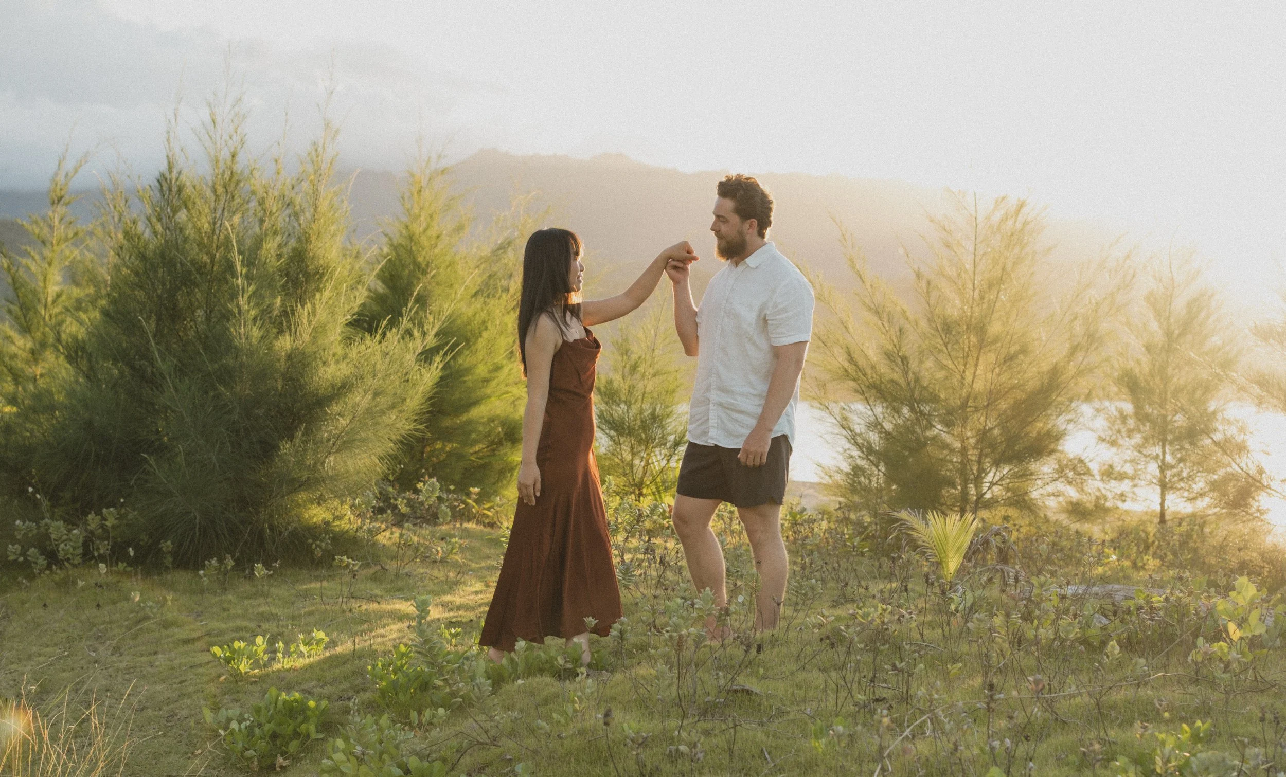 A woman in a brown dress and a man in a white shirt and shorts dancing outdoors among green trees during sunset.