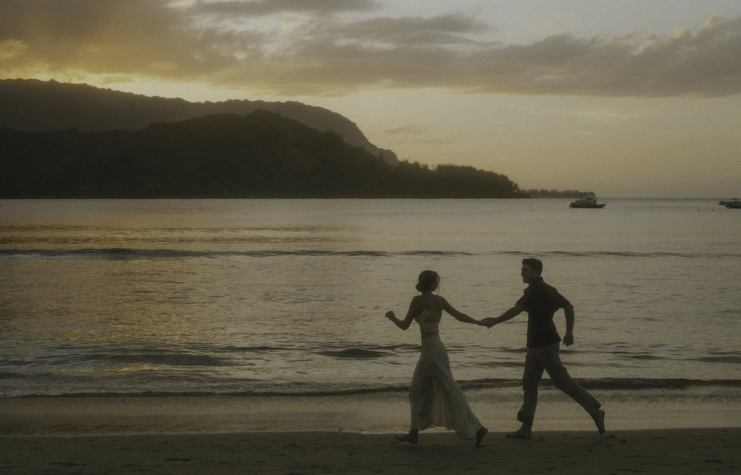 A couple holding hands and walking along the beach at sunset with mountains and boats in the background in Hanalei Bay.
