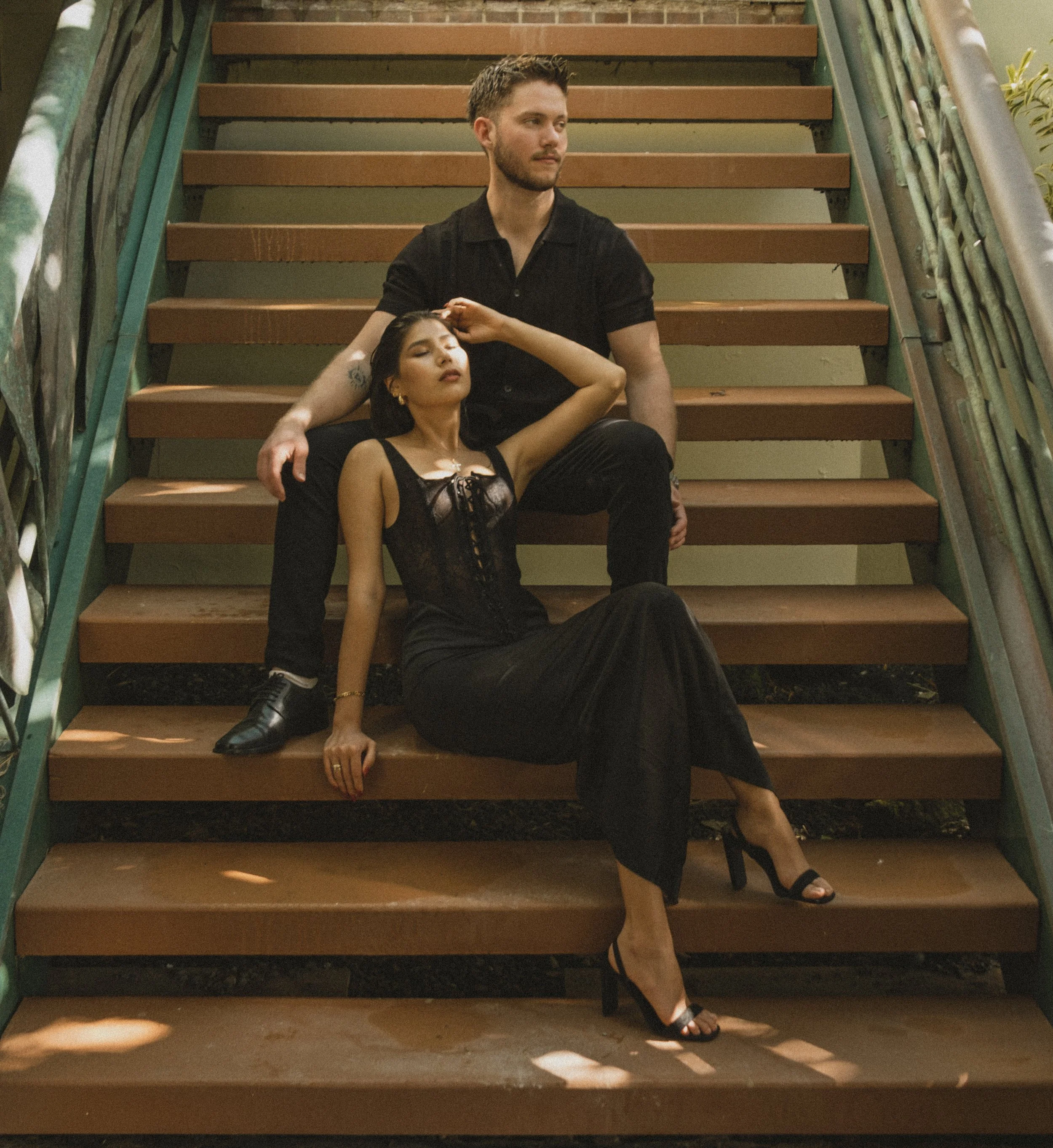 A man and a woman seated on outdoor wooden stairs posing for a photo. The man is sitting with his legs apart, wearing a black shirt and pants. The woman is reclining on the stairs, wearing a black satin dress and high heels, with her eyes closed and