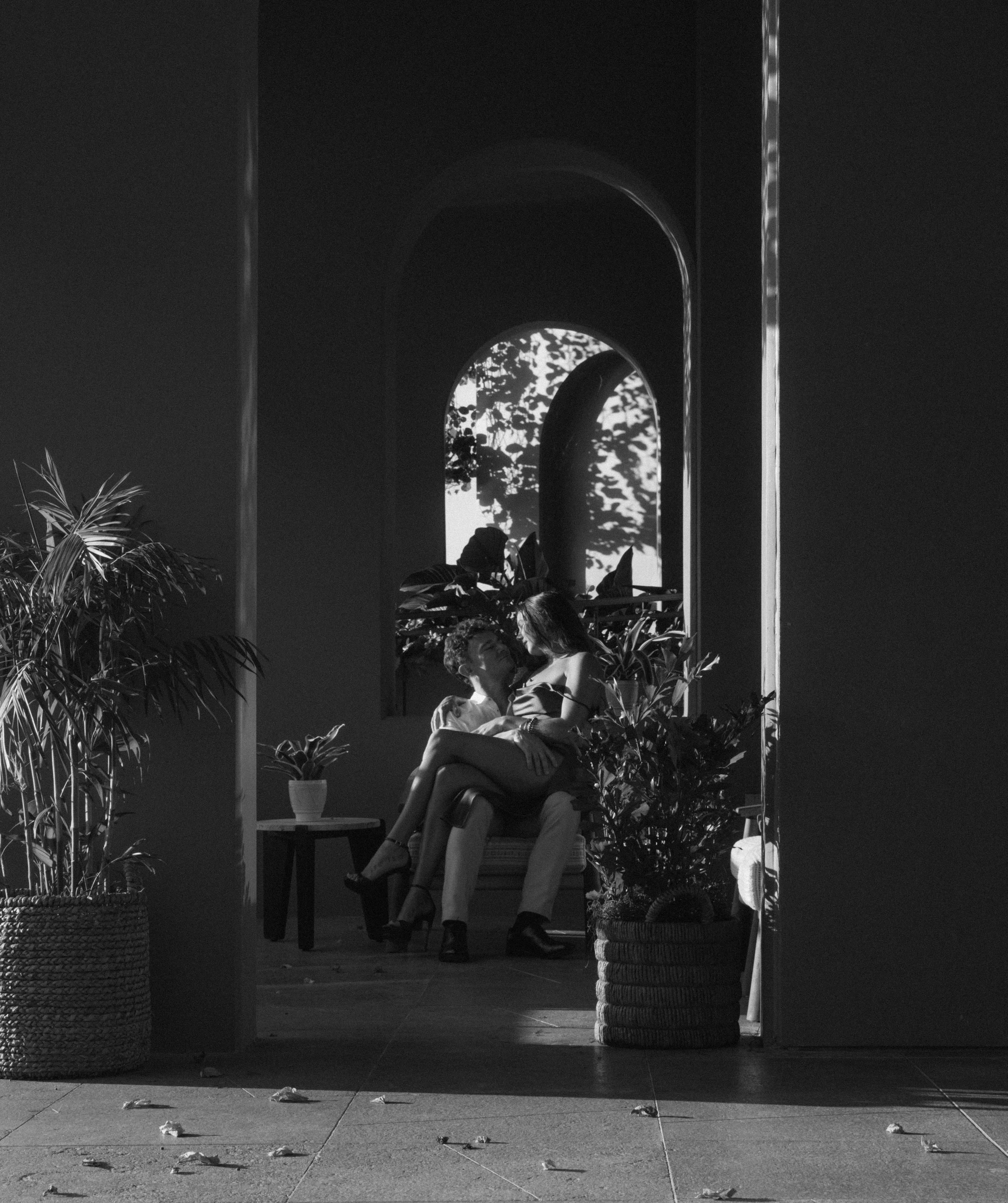Black and white photo of a couple sitting closely on a chair, surrounded by plants, with an arched window and a mirror reflecting foliage in the background.