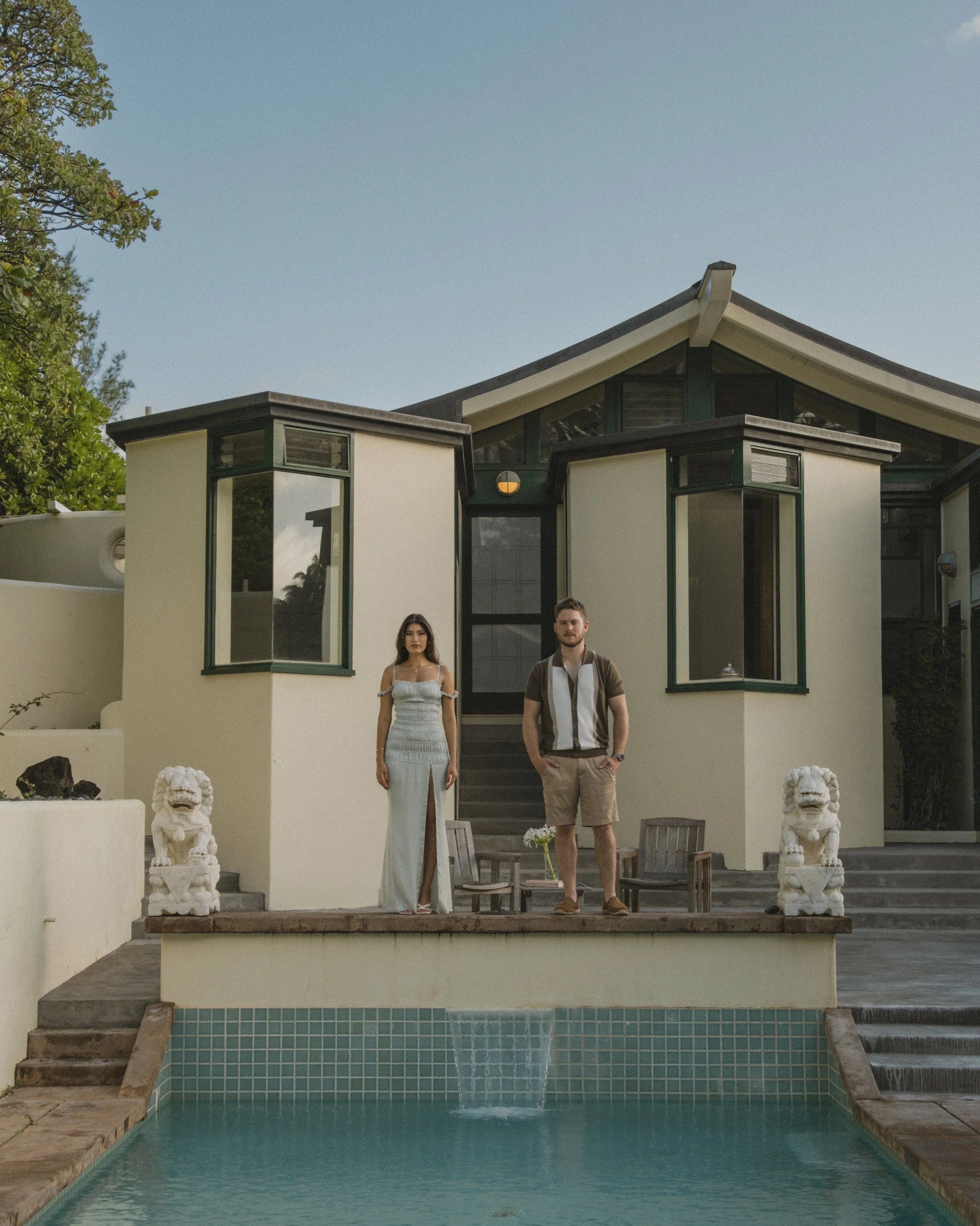 A modern house with a swimming pool and two people standing on the patio. The house has large windows and an architectural design with white walls and dark trim. Lions statues are positioned on either side of the patio.