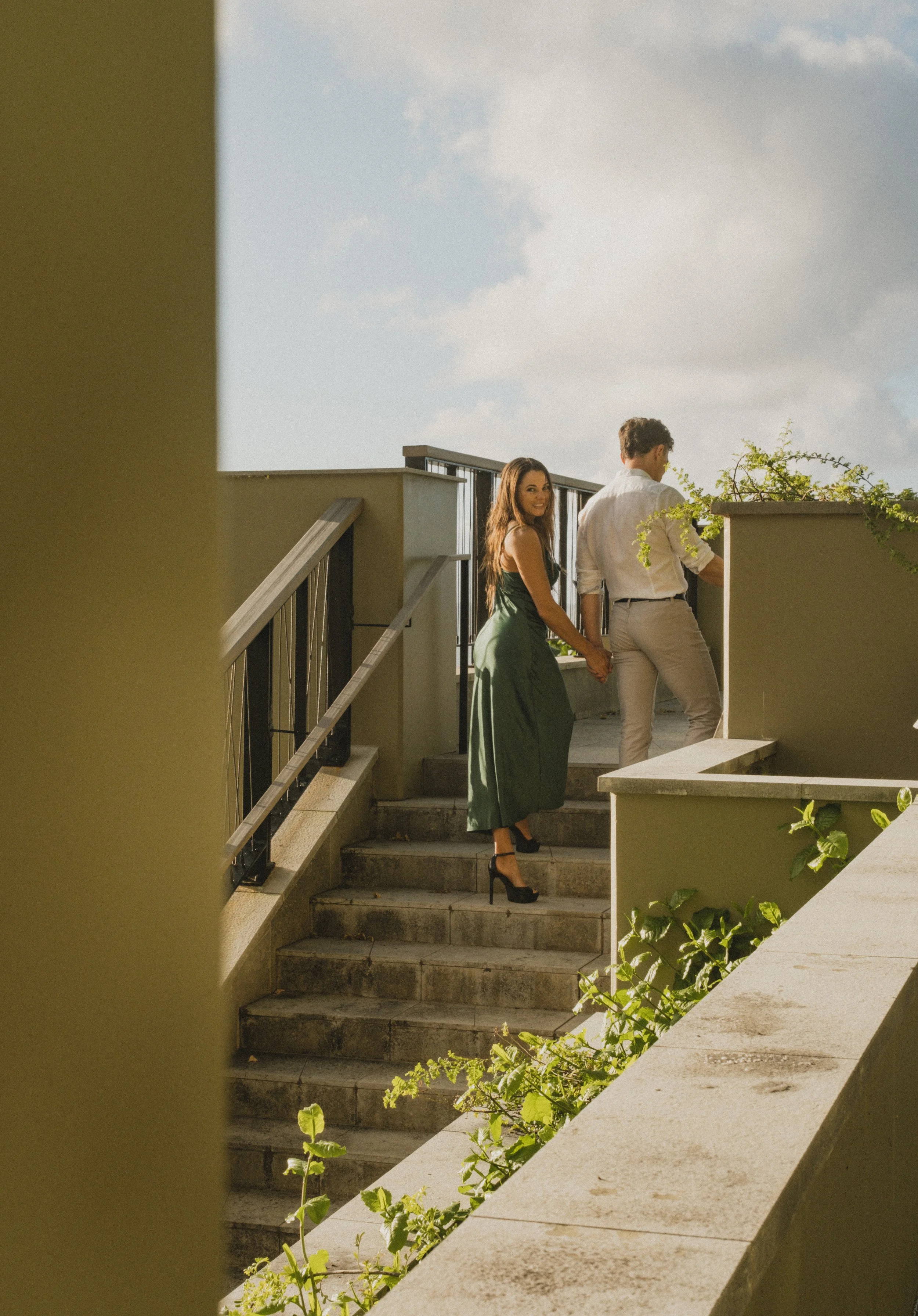 A man and woman holding hands on a staircase on a rooftop, with the woman smiling at the camera, under a bright sky at the 1 Hotel.