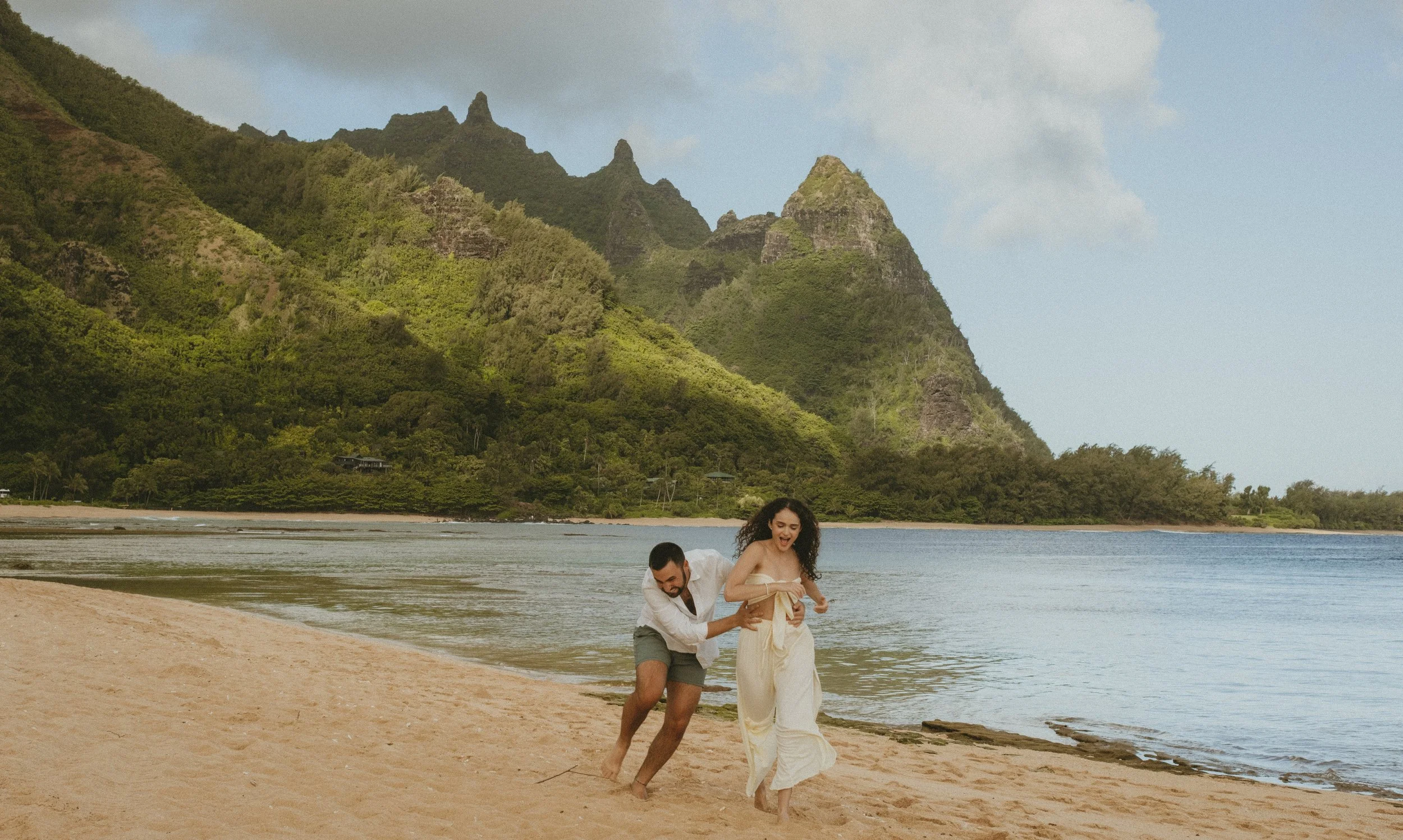 A couple running and playing on sandy beach in Kaua'i with mountains and water in the background.