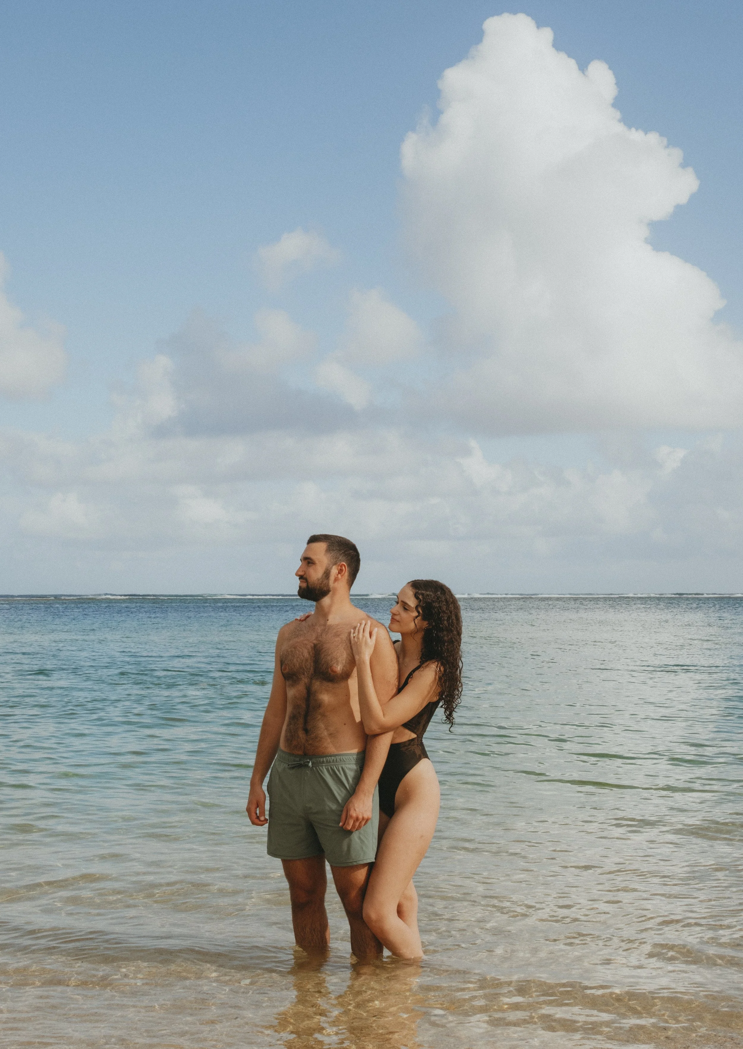 A man and woman standing in the shallow water at a beach in Kaua'i, with the man looking forward and the woman standing closely behind him, touching his shoulder.