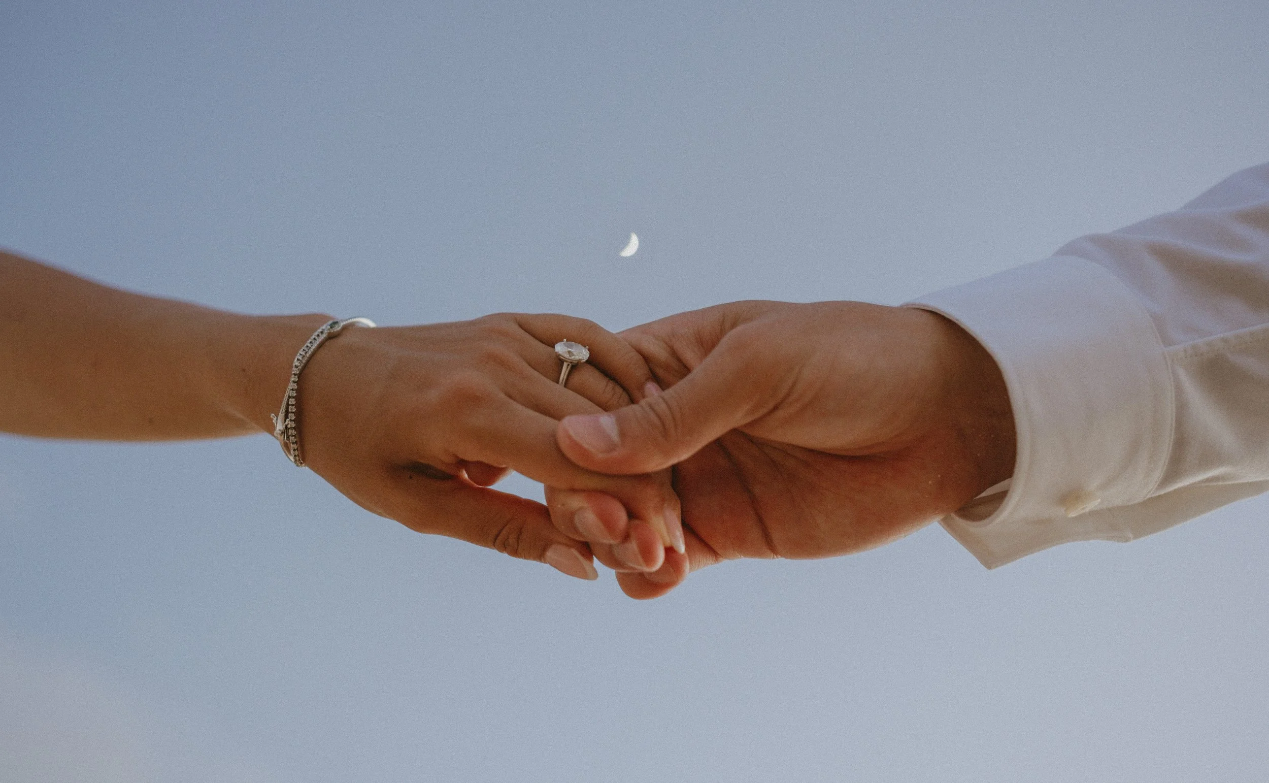 A man and woman holding hands against a blue sky with a crescent moon visible.