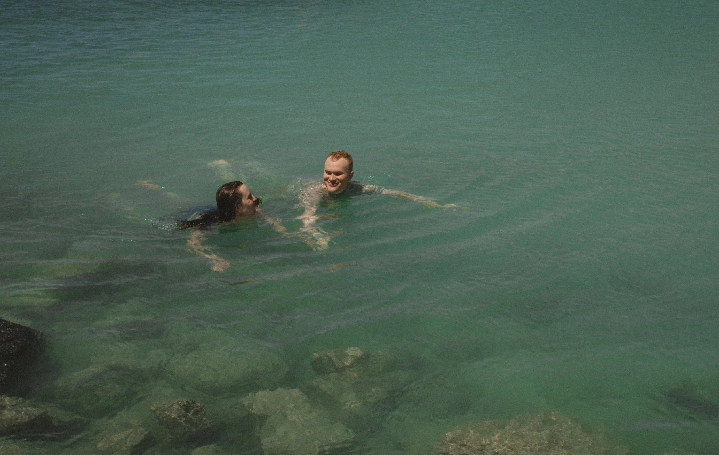 Two people swimming and holding hands in a greenish body of water near some rocks on Big Island, Hawai'i.