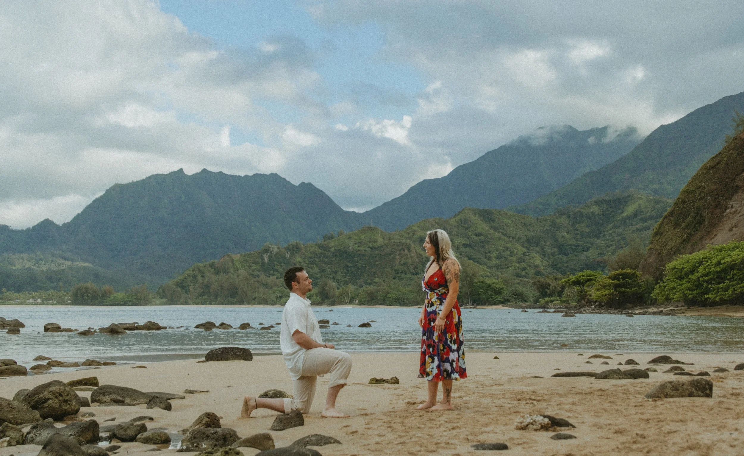 A man is proposing marriage to a woman on a beach in Kaua'i with mountains in the background. The man is kneeling in front of the woman, who is standing, wearing a colorful dress, while he is dressed in a white shirt and beige pants.