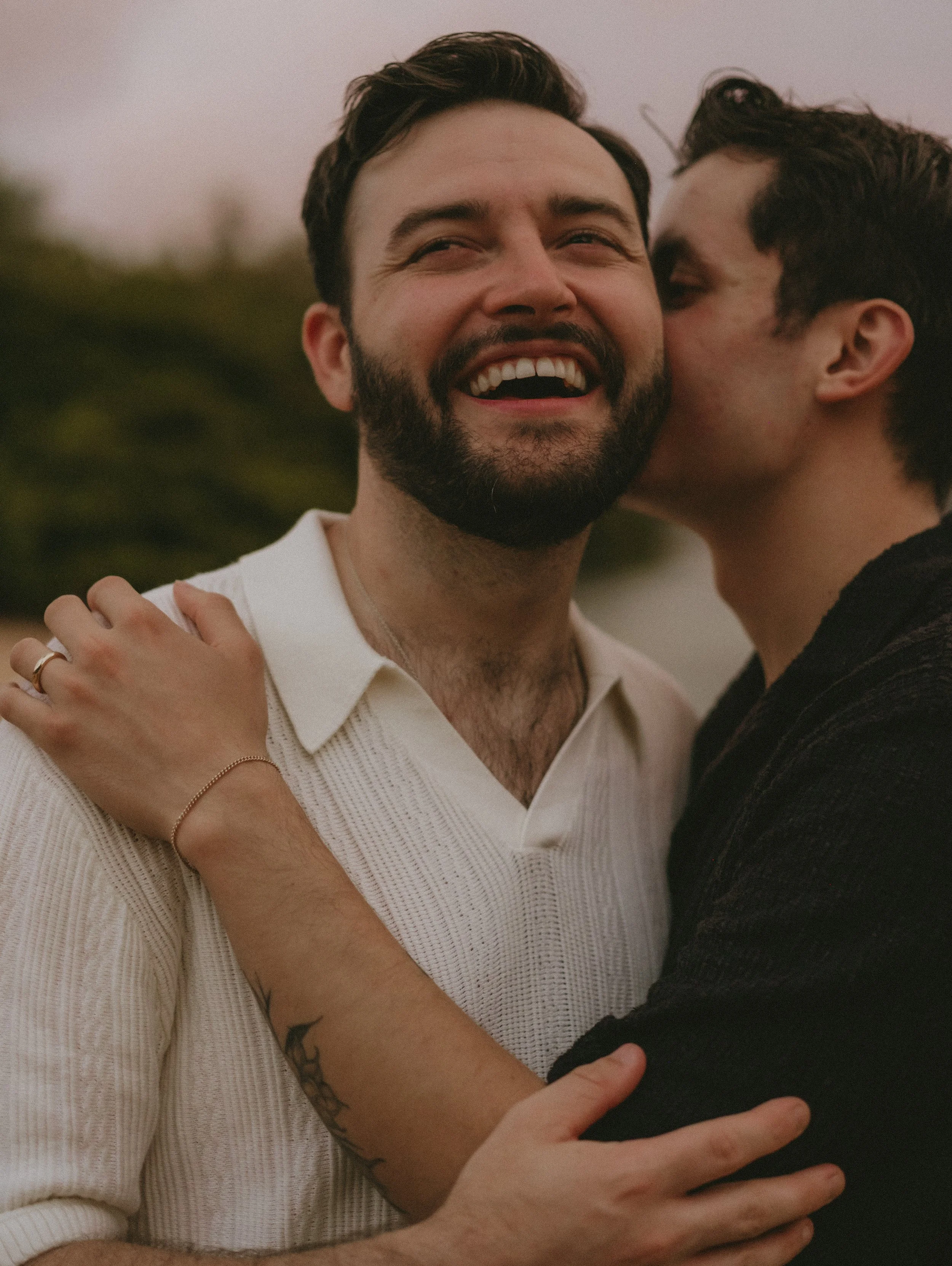 Two men embracing outdoors, with one whispering into the other's ear, smiling and showing affection on Kaua'i.