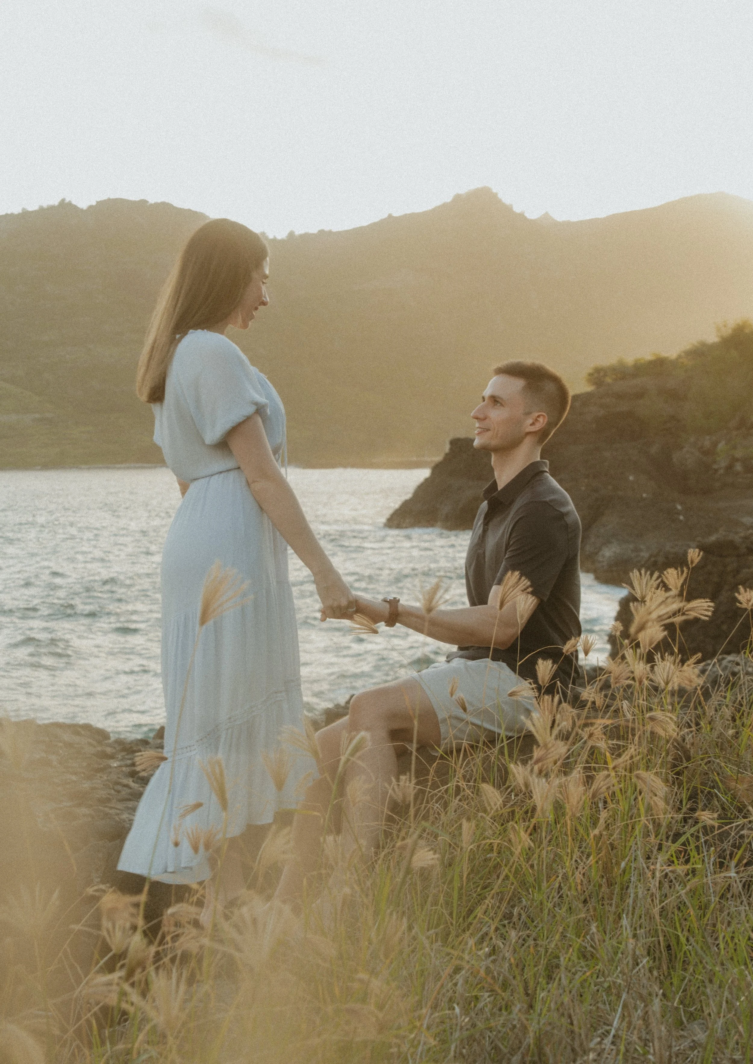 A man proposing to a woman outdoors near the water at sunset, with mountains in the background.