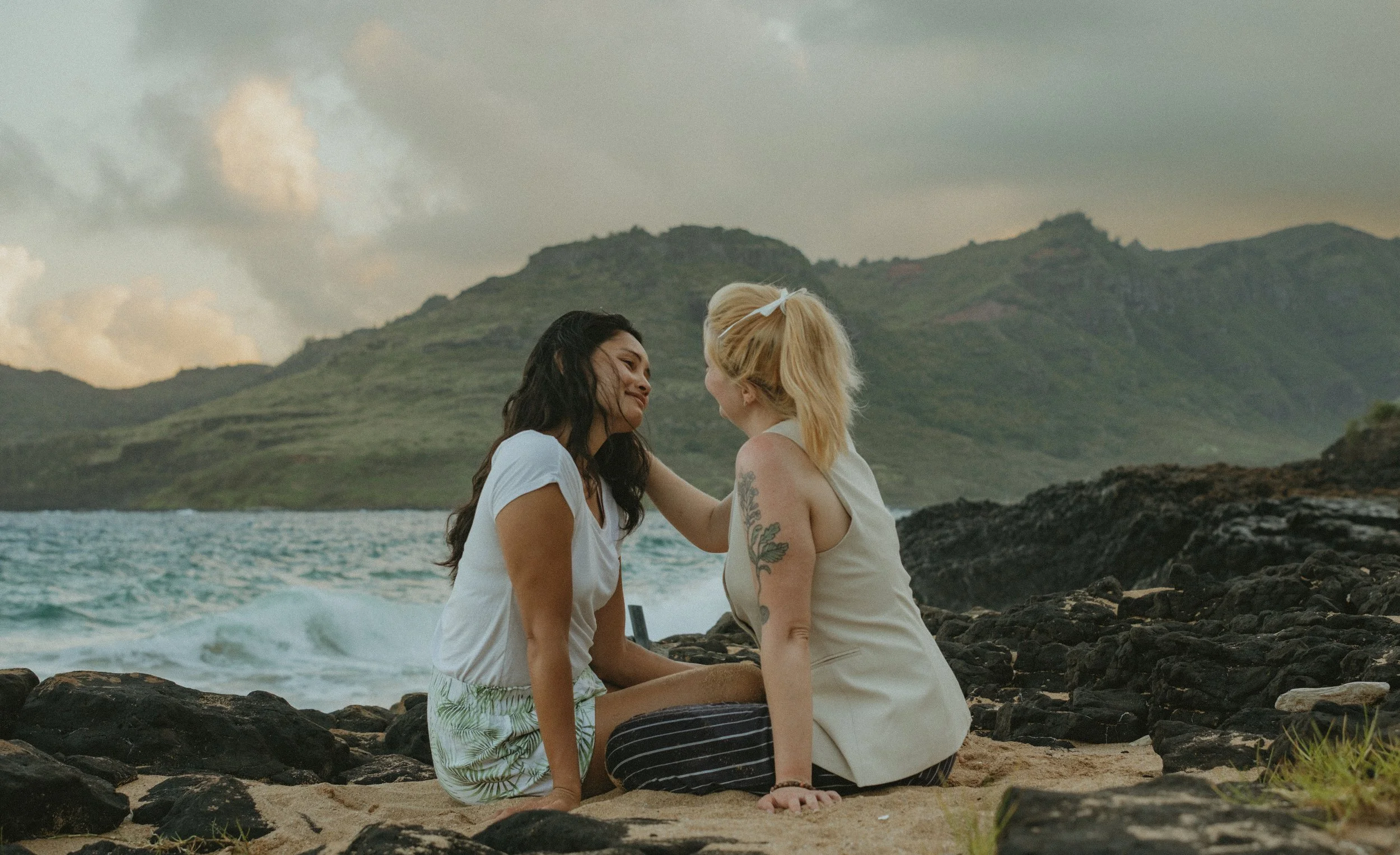 Two women sitting on a rocky beach in Kaua'i, facing each other and smiling, with a green mountainous landscape and cloudy sky in the background.