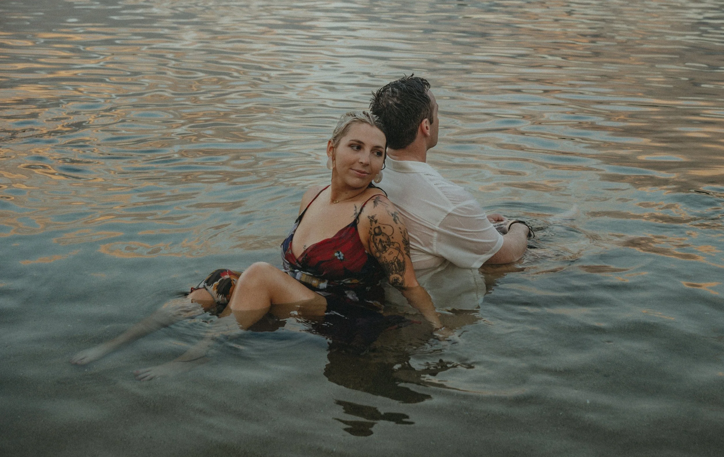 A woman with tattoos sitting on a man in shallow water at sunset, both facing away from the camera. On Kaua'i