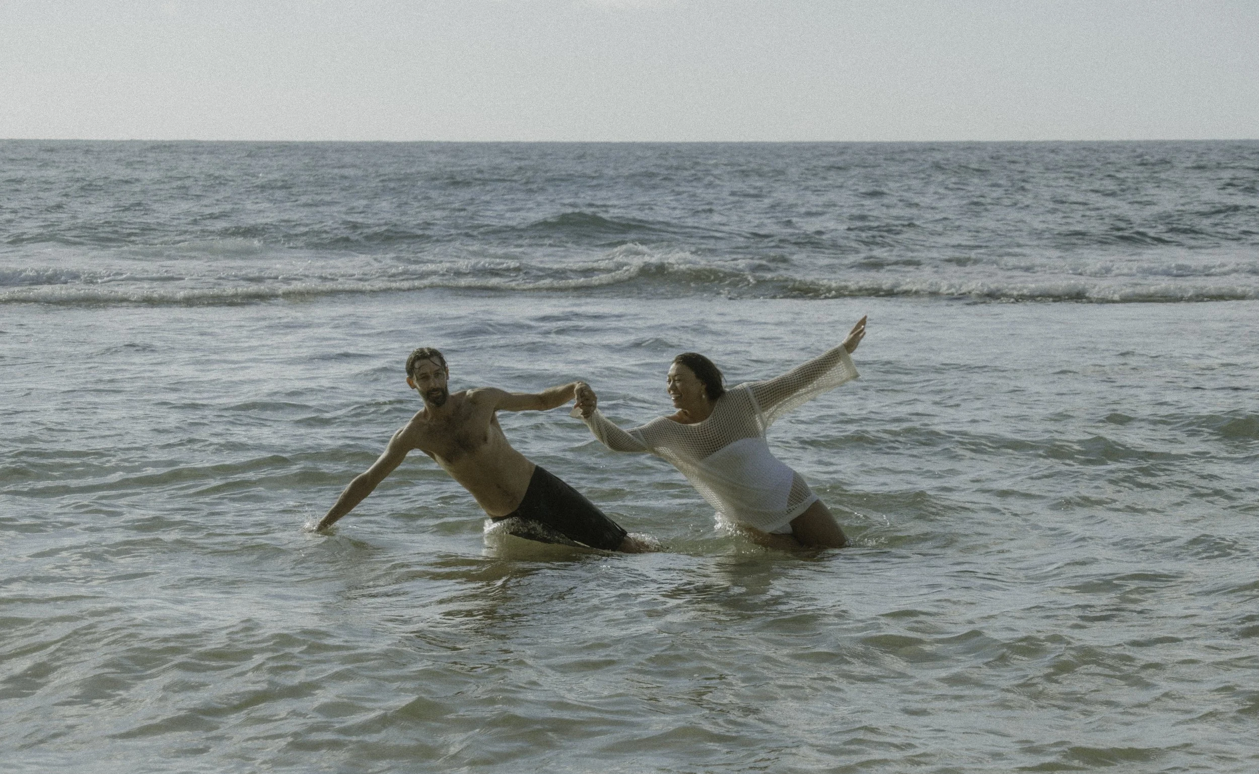 A man and a woman are playing and holding hands in the shallow ocean water on a beach. The man is shirtless and wearing dark swim trunks, while the woman is wearing a white mesh cover-up over swimwear. They appear to be having fun, smiling and enjoyi