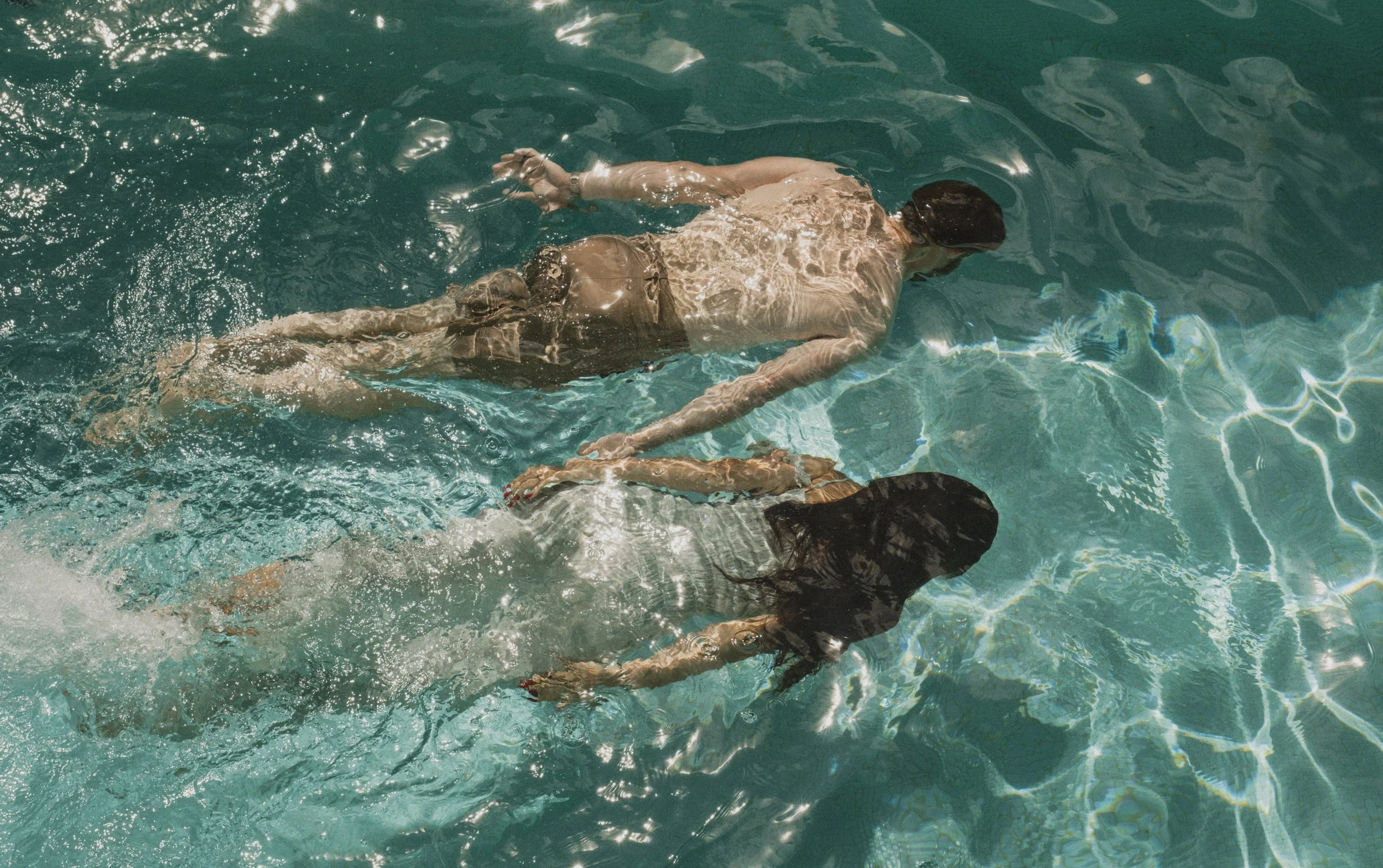 Two women are swimming in a pool, holding hands, with sunlight reflecting on the water surface.