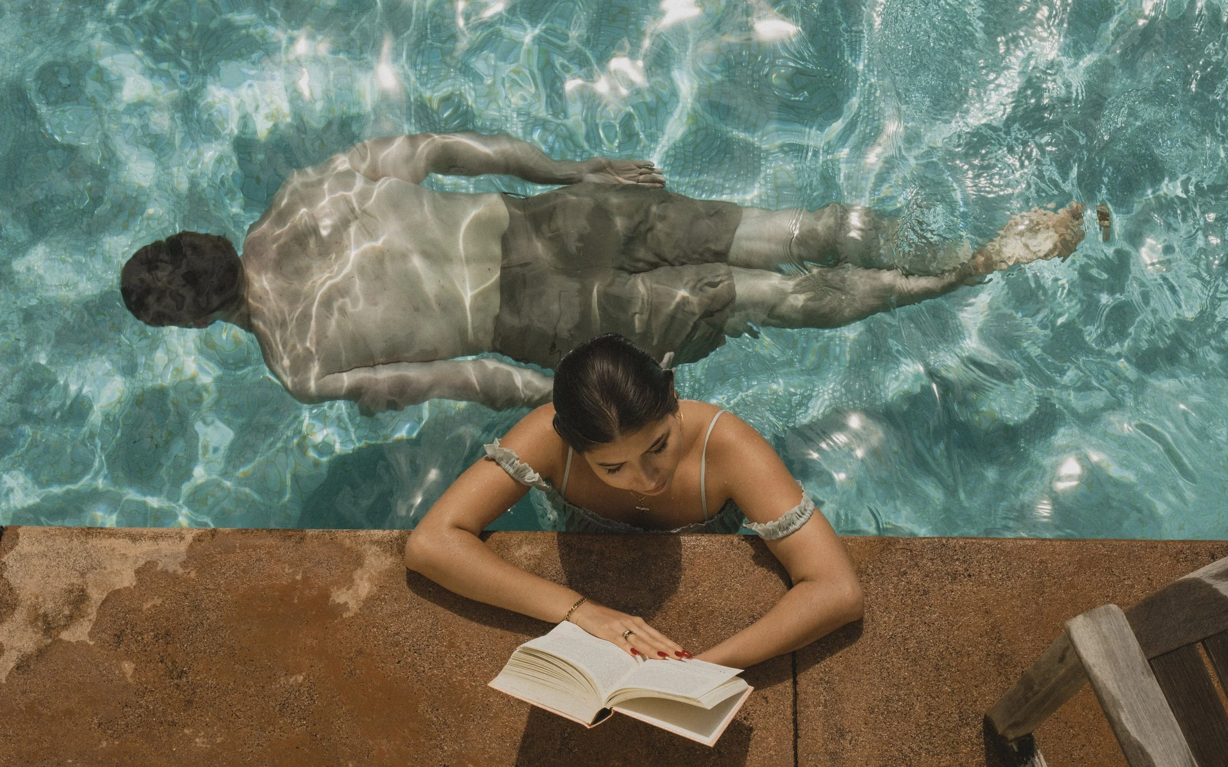 A woman with dark hair reading a book while leaning on the edge of a swimming pool, with her legs submerged in the water, as a shirtless man lies floating in the pool's water nearby.