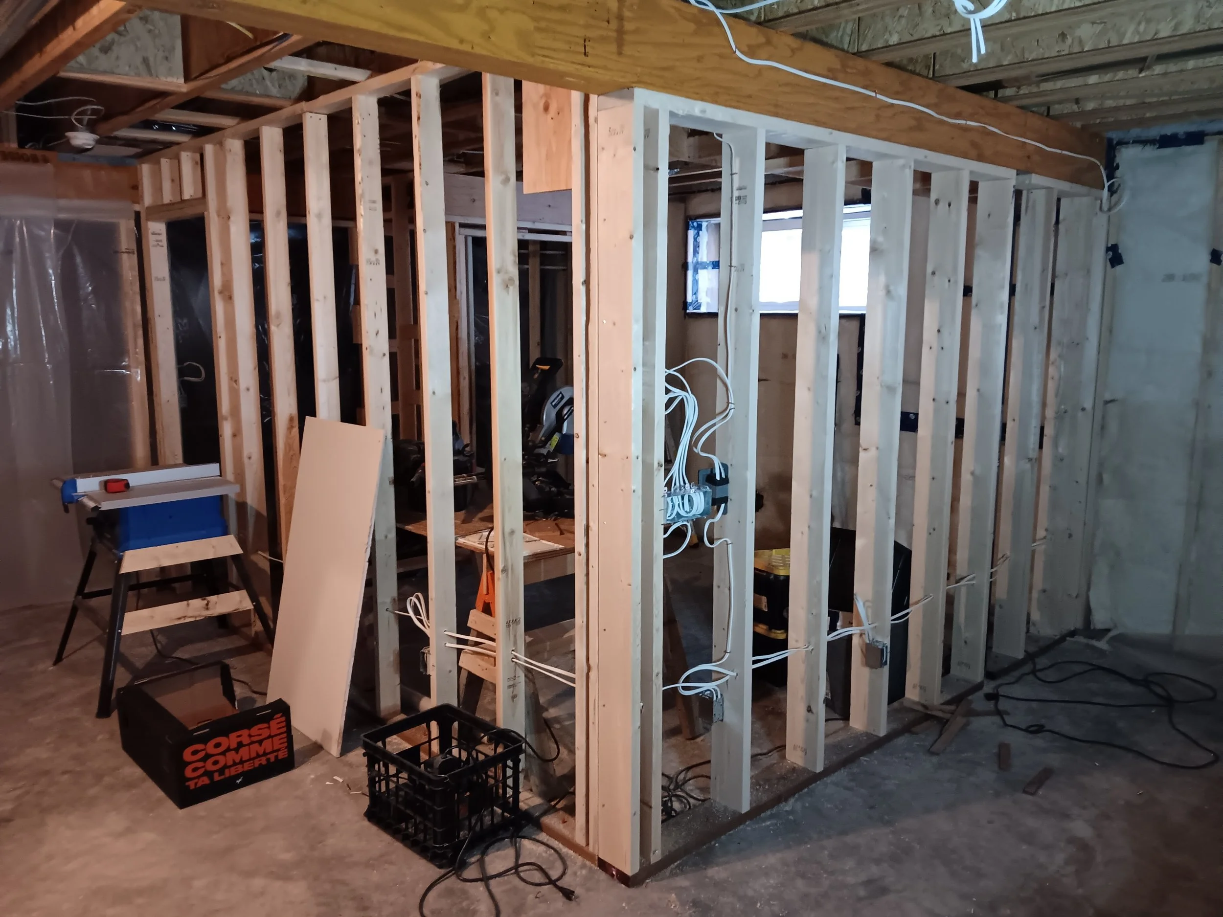 Construction framing of interior wall with wooden studs, electrical wiring, and tools in a basement or utility room