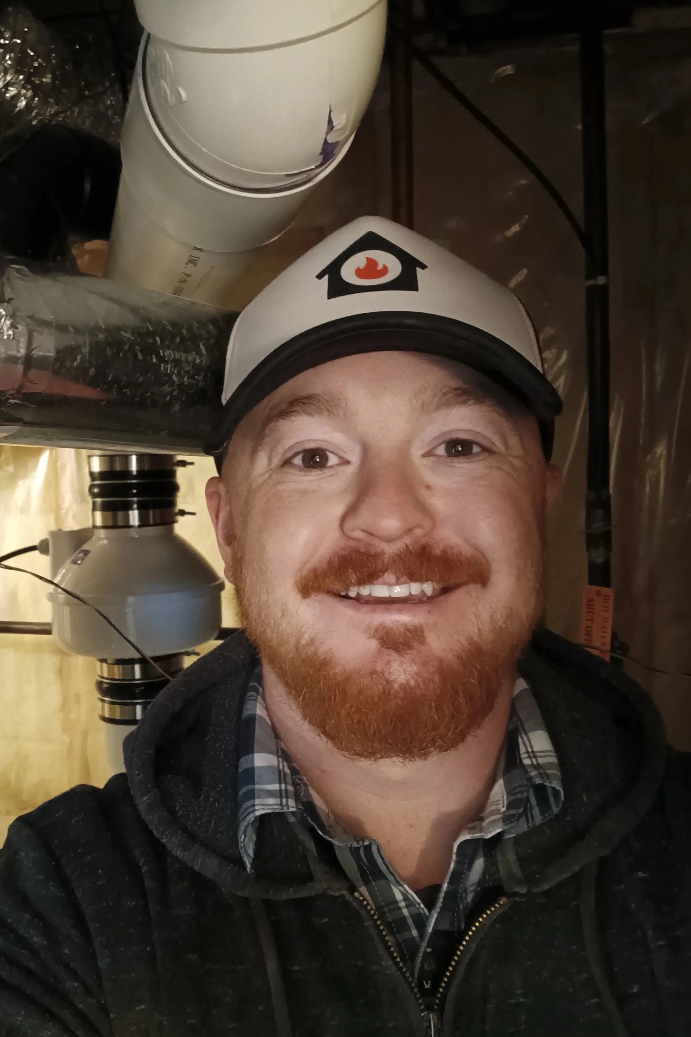 A man with a red beard and mustache smiling, wearing a white baseball cap with a house and flame logo, plaid shirt, and black hoodie, standing in an industrial or mechanical environment with pipes and machinery in the background.