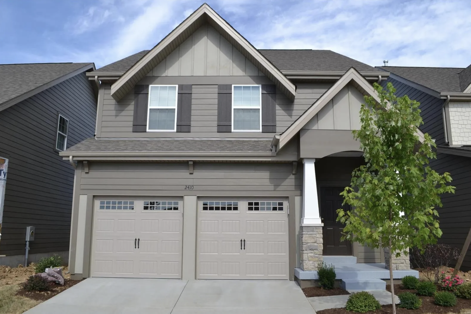 A modern two-story house with gray siding, a tiled roof, and a double garage with decorative windows. There is a green tree in front and some shrubs, with a small set of stairs leading to the front door.