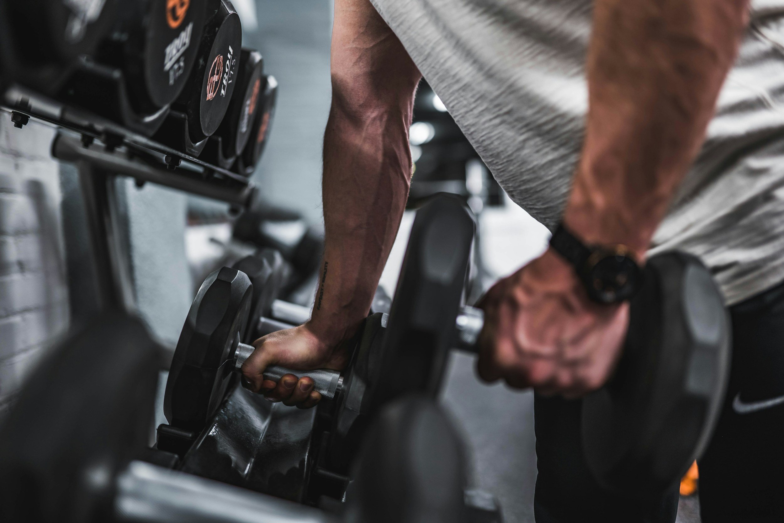 A person bending over and gripping a dumbbell during a weightlifting workout in a gym. The image focuses on the person's arm muscles and the dumbbell, with gym equipment visible in the background.