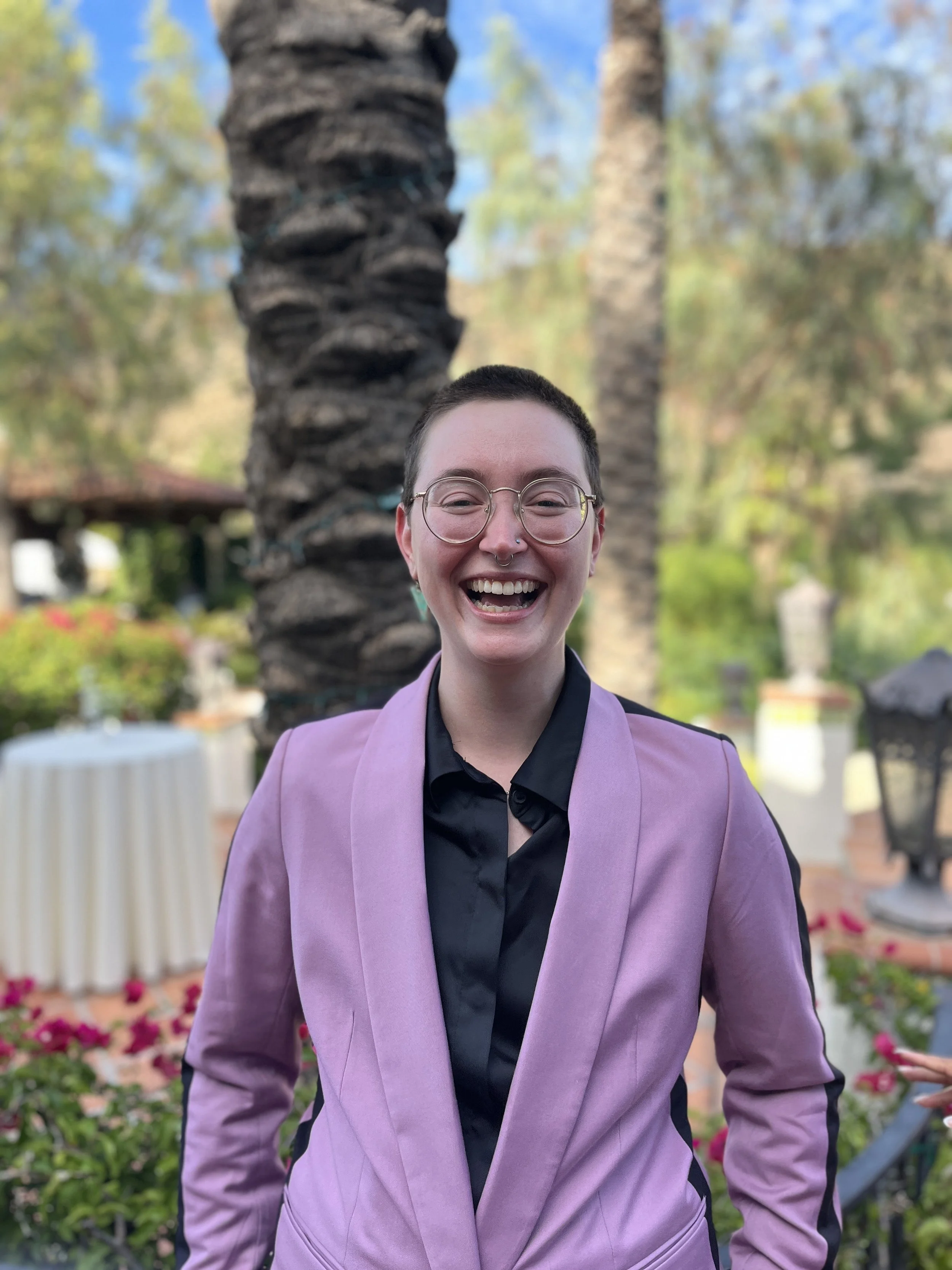 A person with short, brown hair and wire-rimmed glasses smiles at the camera in a lavender suit jacket.
