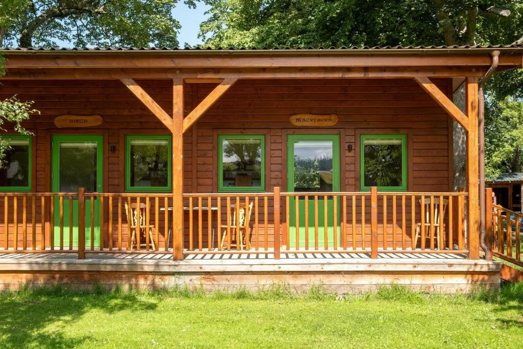 Wooden cabin with a porch, green window frames, and benches, surrounded by green grass and trees. Marcassie Farm, Scotland