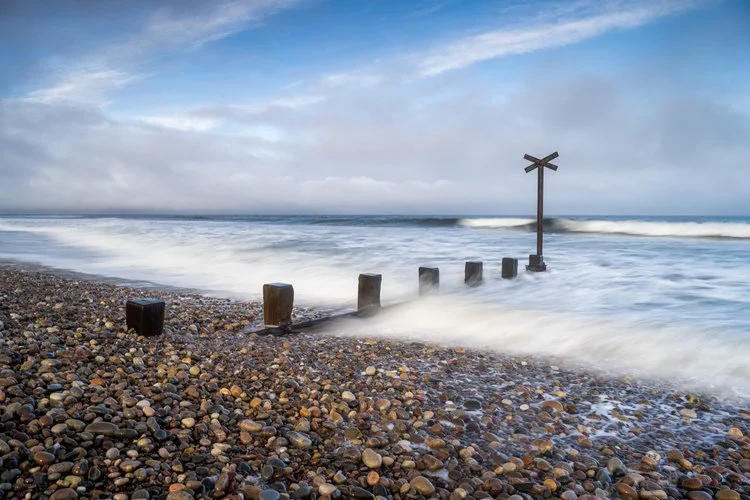 A pebble beach with a series of wooden posts and a signpost near the water's edge, with waves and a cloudy sky in the background. Findhorn Beach landscape photography