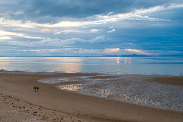 Two people walking on a sandy beach near calm water, with a cloudy sky and a distant landmass under a partly bright sky