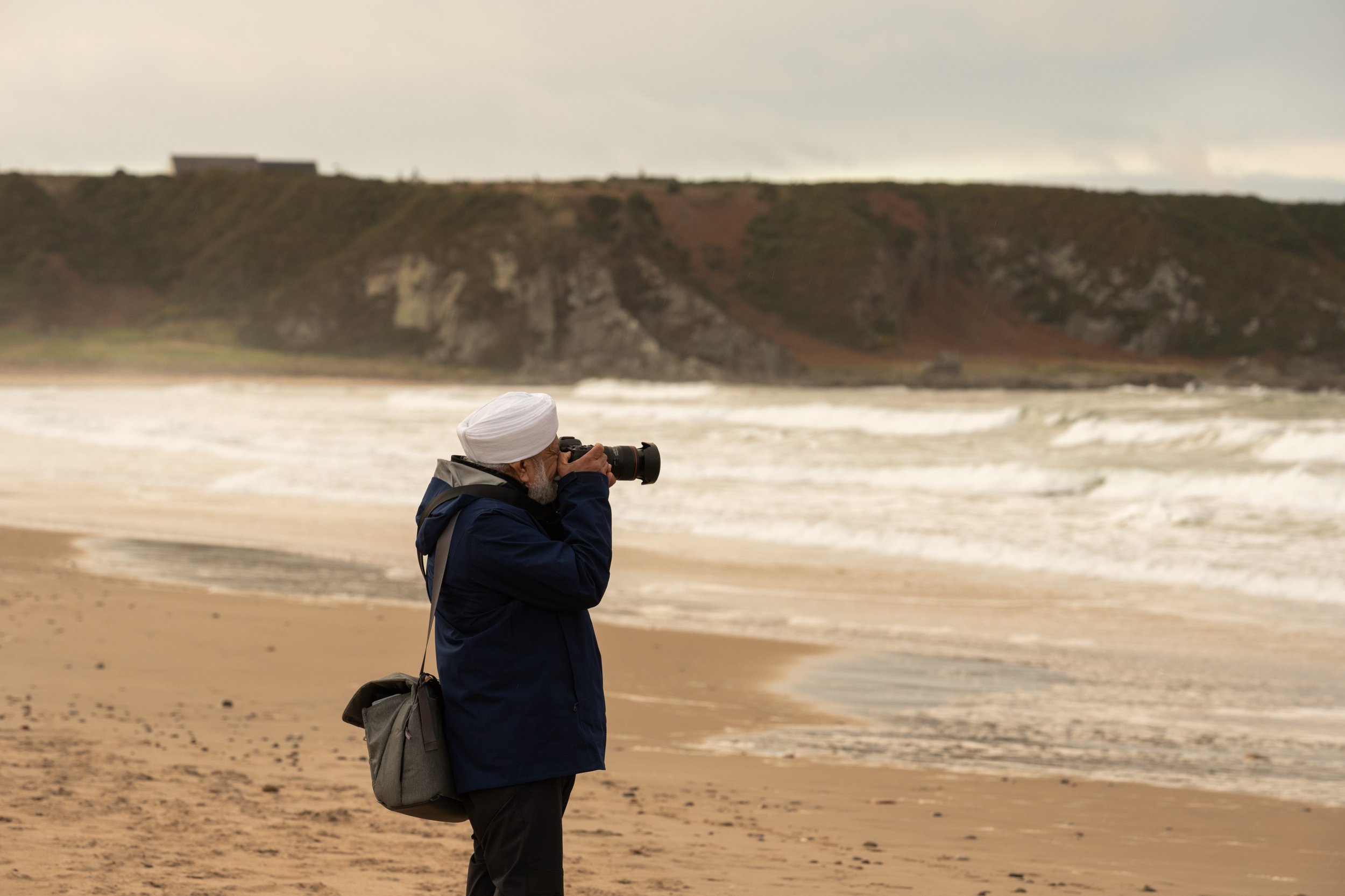 A man wearing a white turban and dark jacket standing on a sandy beach, taking a photograph with a camera. The ocean waves are in the background along with cliffs and a cloudy sky.