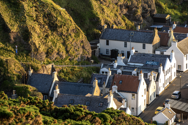 A cluster of white houses with chimneys, situated at the base of a hill with greenery, some houses have grey roofs and others have red roofs, and one building has a sign that reads 'HOTEL'. Pennan, Aberdeenshire