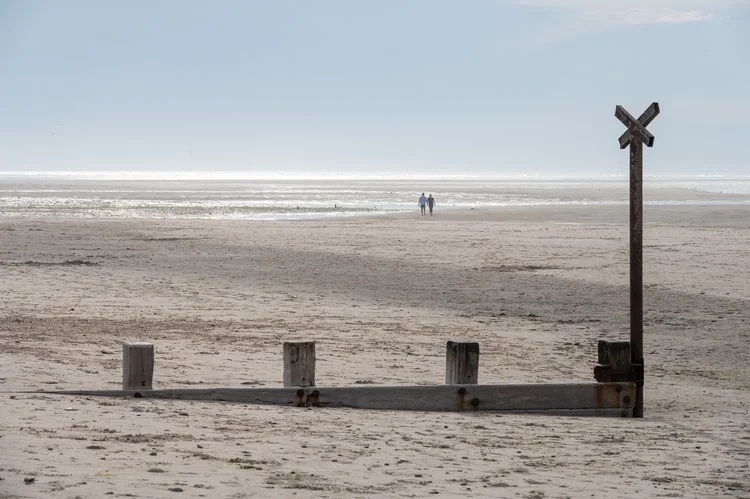 Empty sandy beach with a wooden railroad crossing sign and posts, two people walking in the distance by the water. Findhorn Beach, Scotland