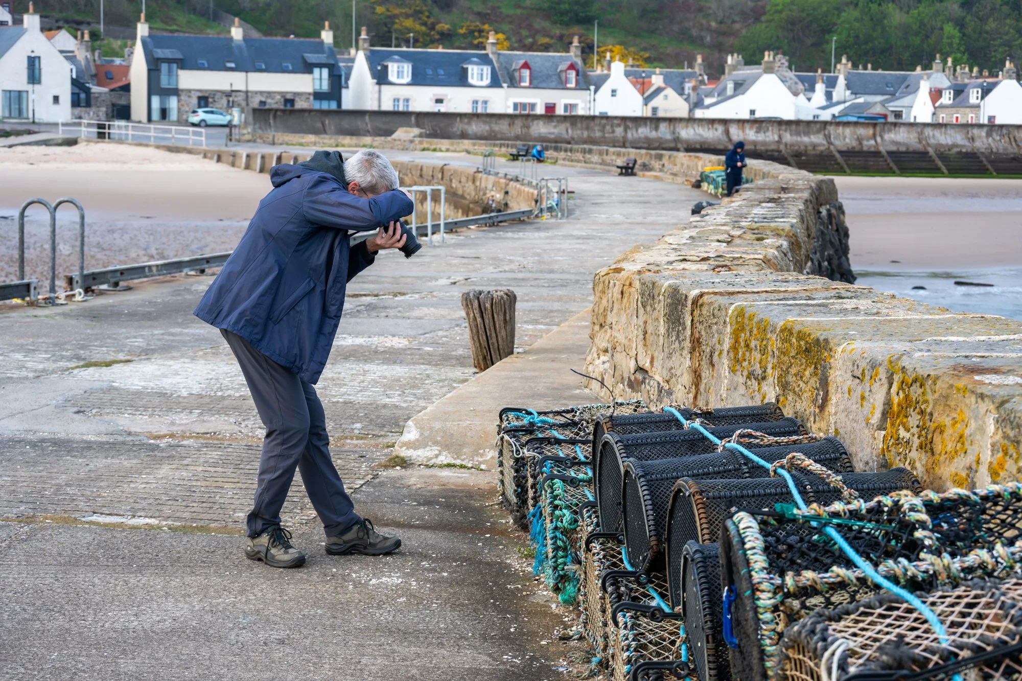 A person wearing a dark blue jacket  taking a photo with a camera on Cullen pier, with a row of lobster traps stacked along the stone wall in the foreground during mindful photography workshop