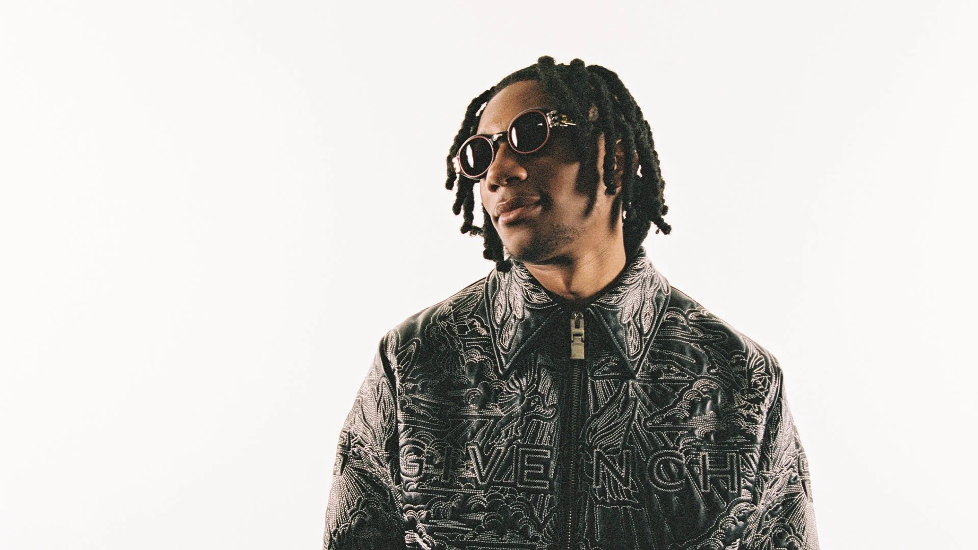A person with shoulder-length dreadlocks wearing round sunglasses and a black and silver embroidered jacket with intricate patterns and the word "GIVENCHY" on the front, standing against a plain white background.