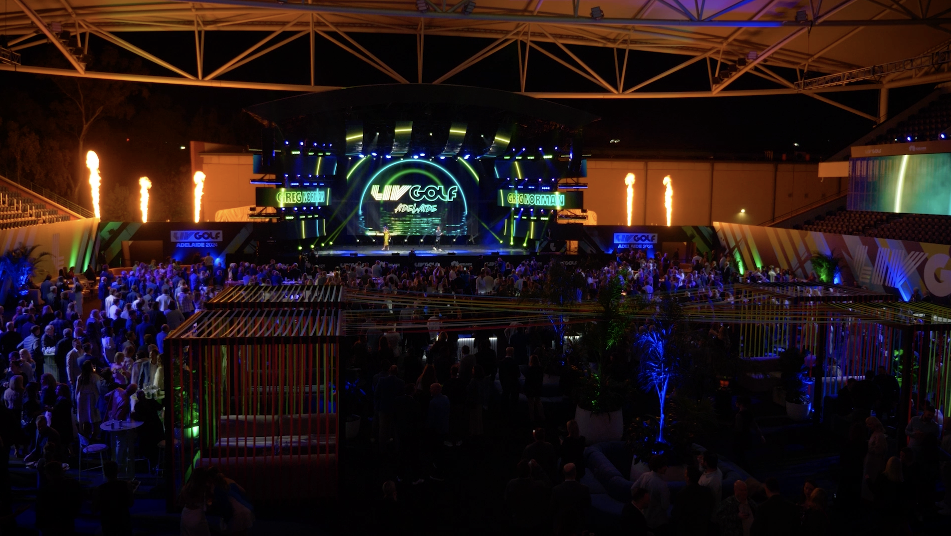 Nighttime scene at LIV Golf Adelaide 2023, with a stage featuring bright neon lights, screens displaying LIV GOLF ADELAIDE, and fire pyrotechnics, surrounded by a large crowd of attendees.