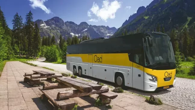 Een grote bus geparkeerd in een bergachtig landschap met groene bomen en een heldere blauwe hemel.
