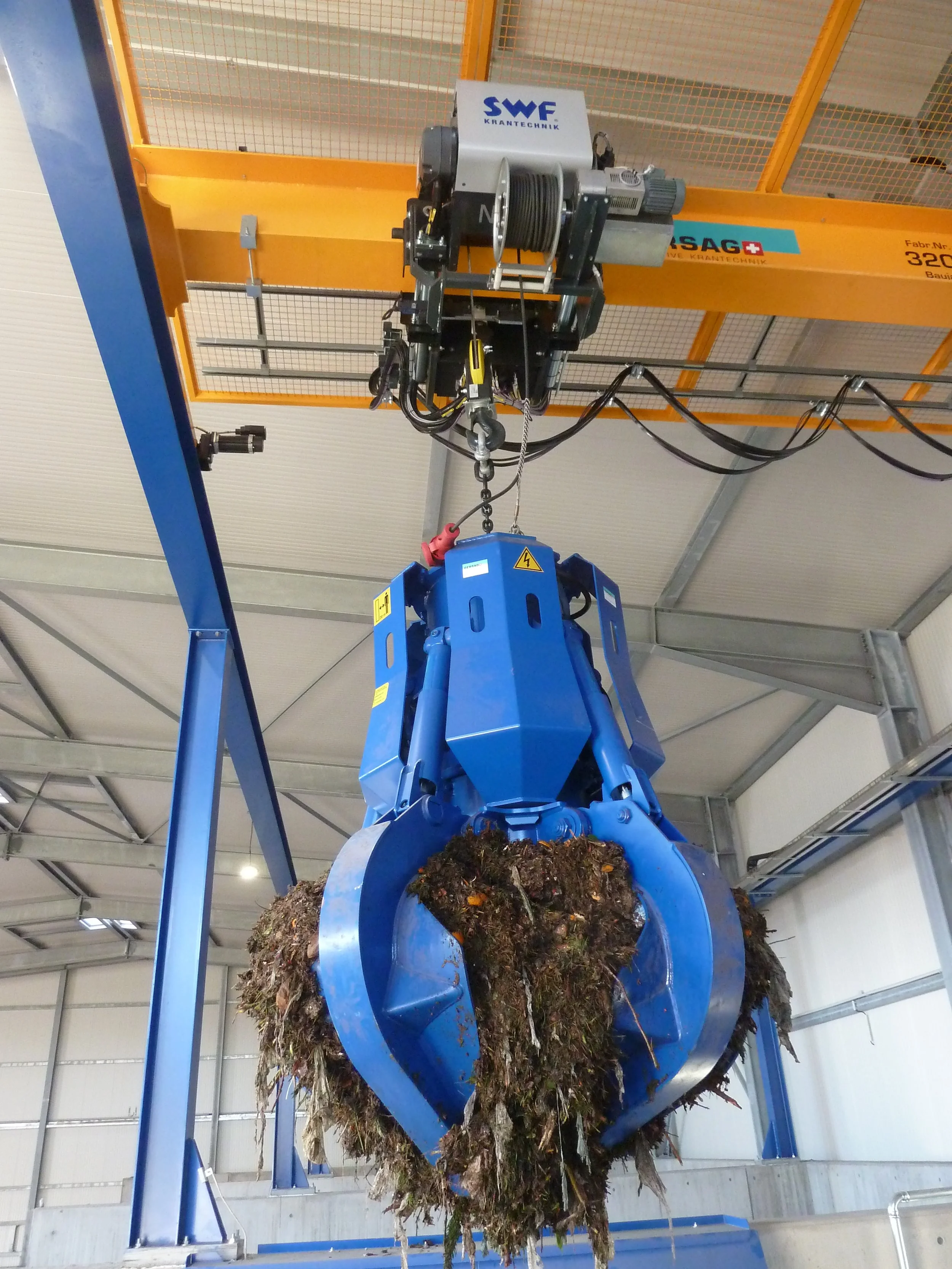 A large electrohydraulic grab is lifting organic waste inside a warehouse.