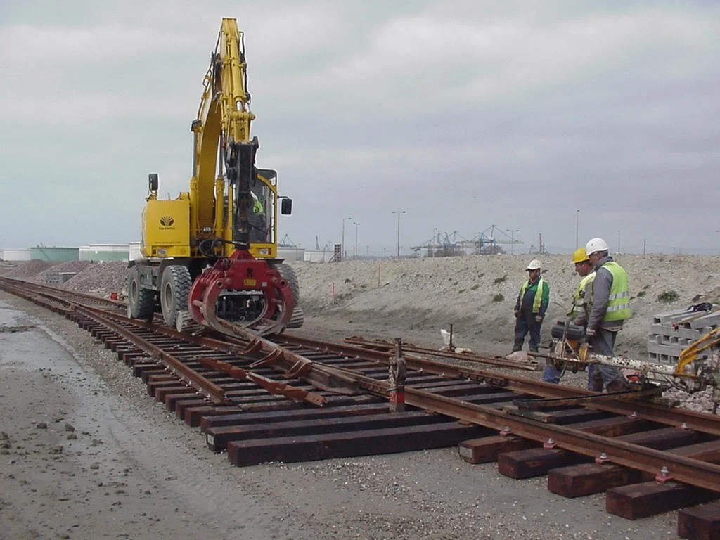 A yellow machine on railway tracks using a Rozzi clamshell to load gravel.