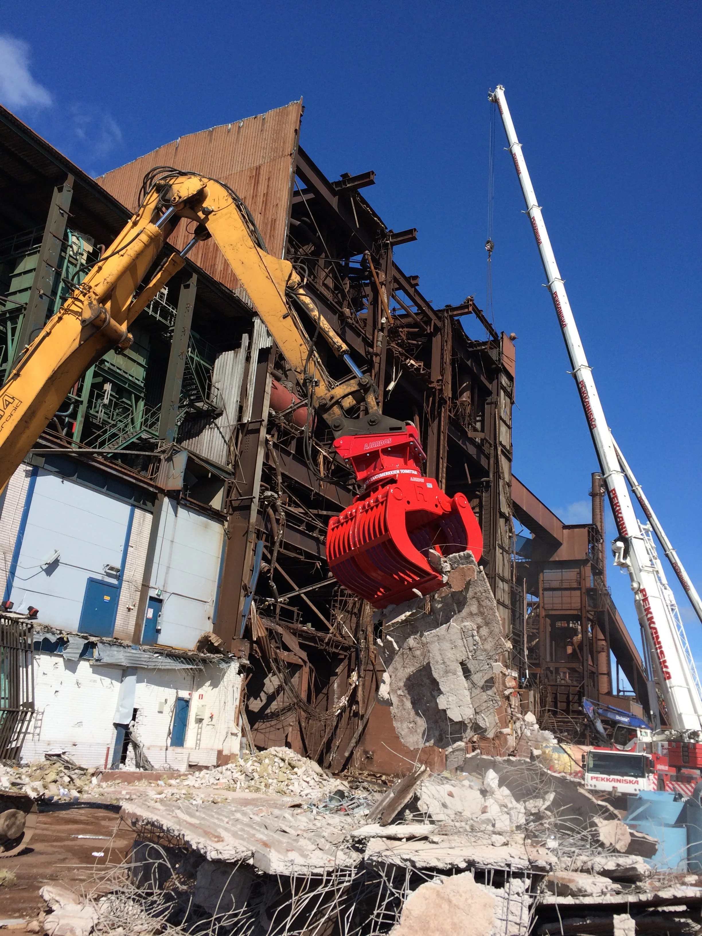 Construction site with a building partially demolished, heavy machinery removing debris, and a large crane next to the site. Clear blue sky in the background.