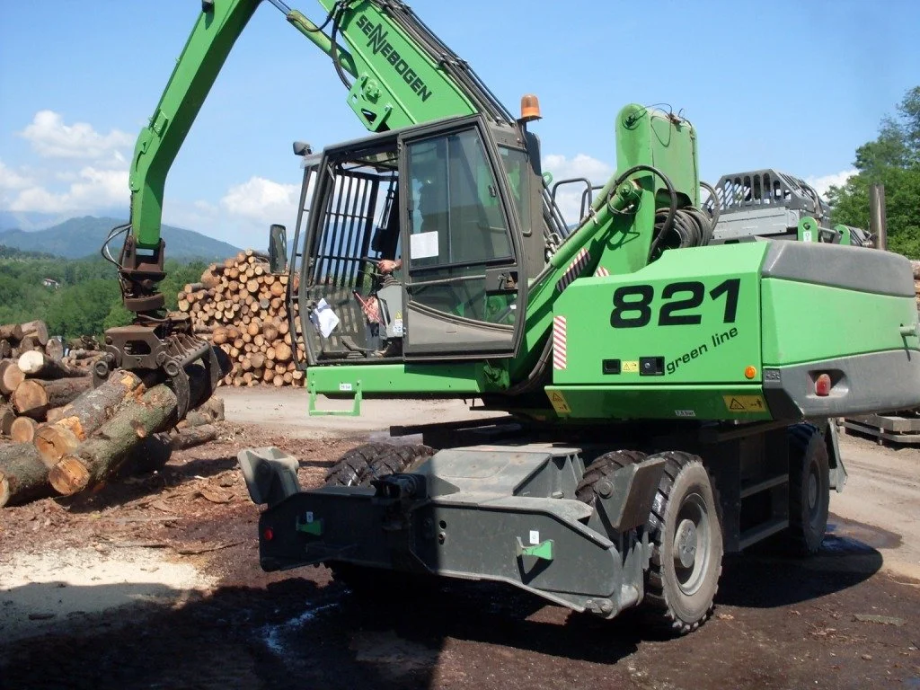 A green Senebogen 821 excavator stacking logs at a lumber yard against a backdrop of mountains and blue sky.