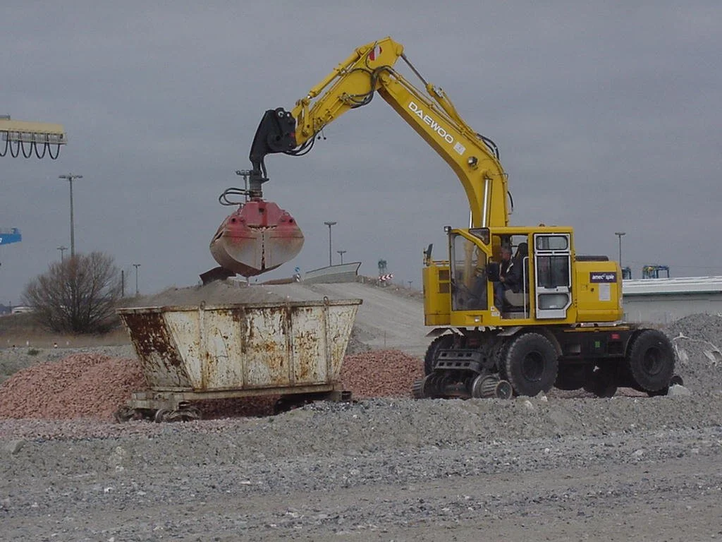 A yellow machine on railway tracks using a Rozzi clamshell to load gravel.
