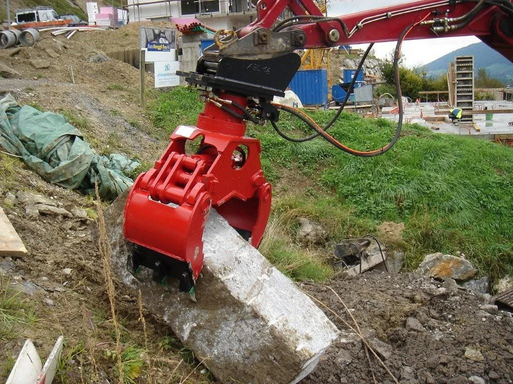 Close-up of a construction excavator's hydraulic grapple attachment holding a large concrete pipe segment on a construction site with greenery and buildings in the background.