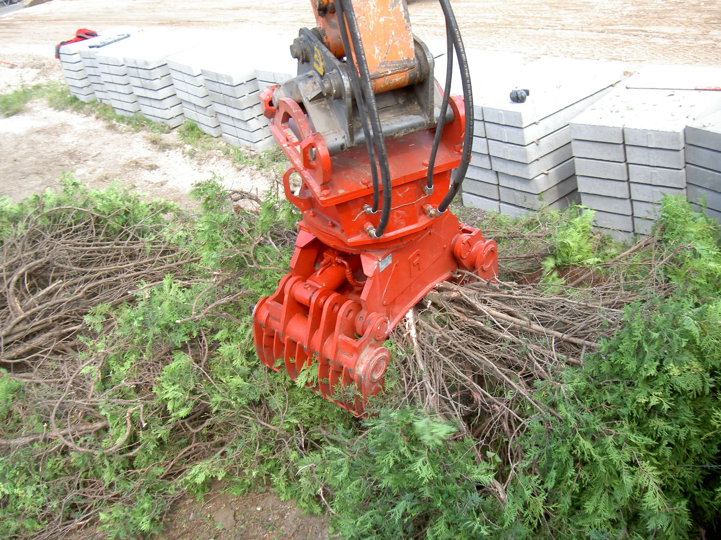 A hydraulic log splitter with a red base and orange hydraulic arm, positioned in a forest area with green foliage and cut branches in the foreground, and a background of tiled concrete blocks and ground.