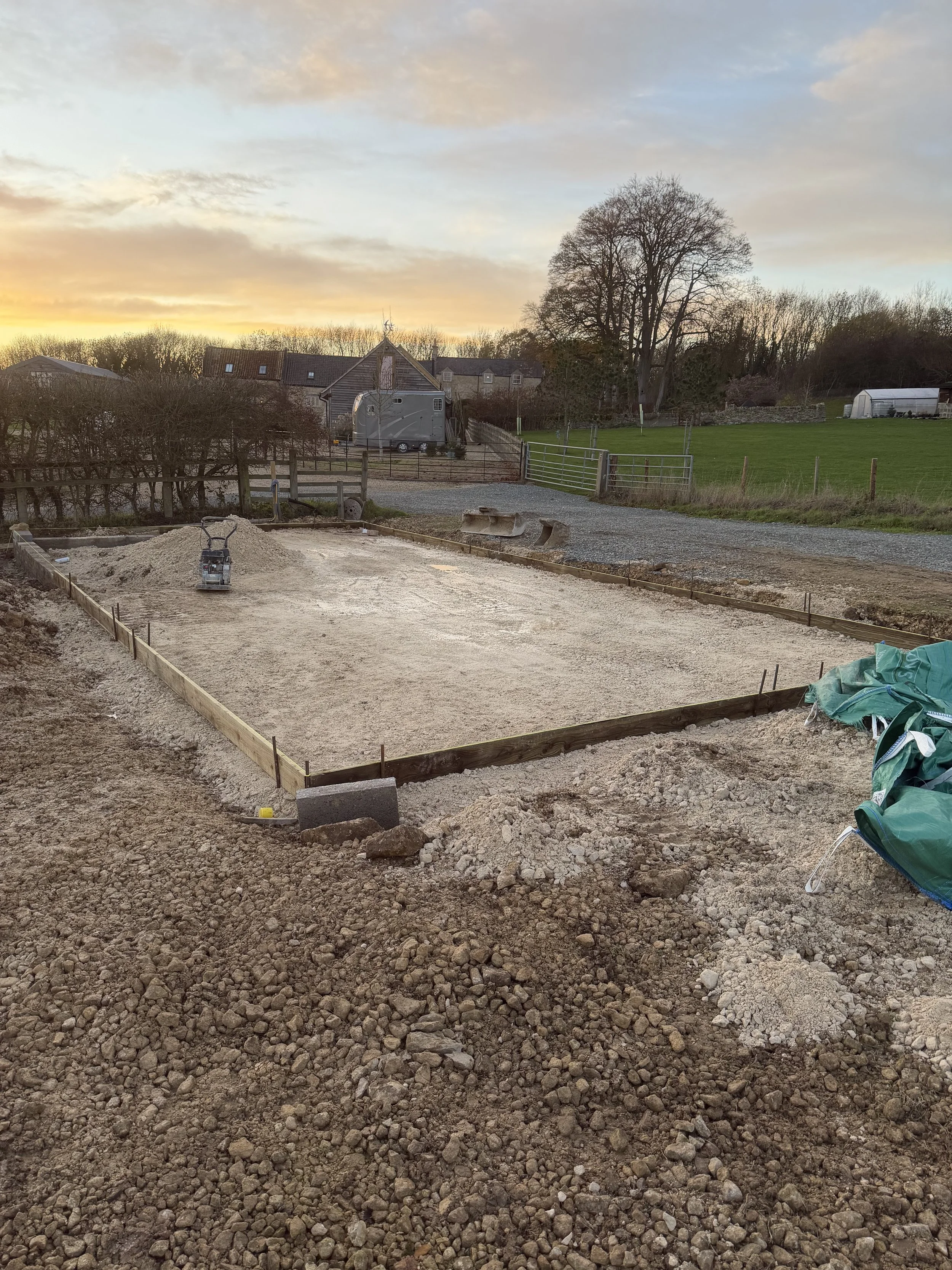A construction site at sunset with a partially built foundation, wood framing, and gravel path. In the background, there are trees, houses, a shed, and a trailer, with a sunset sky overhead.