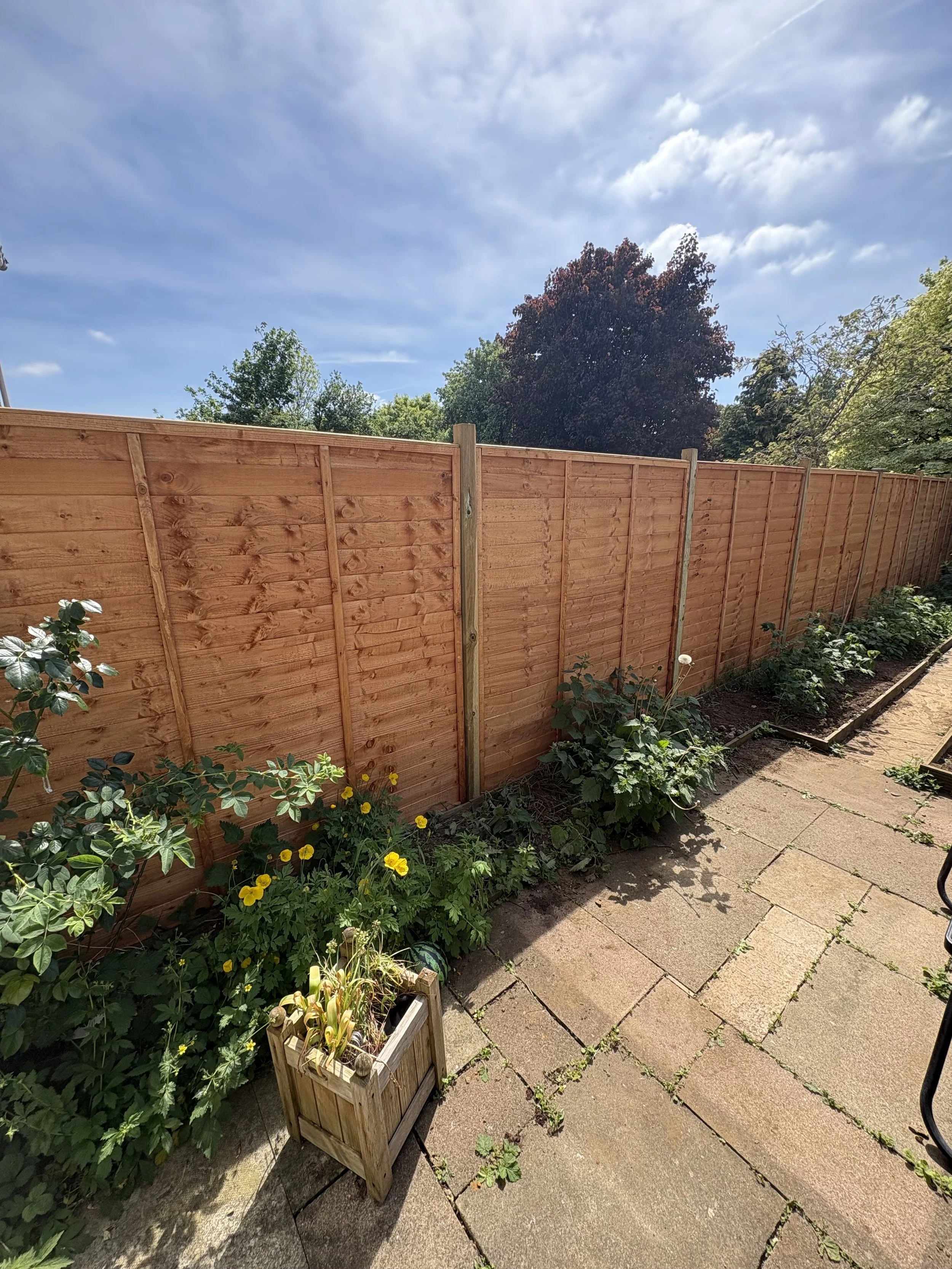 A backyard garden with a wooden fence, flowering plants, and a paved patio area.
