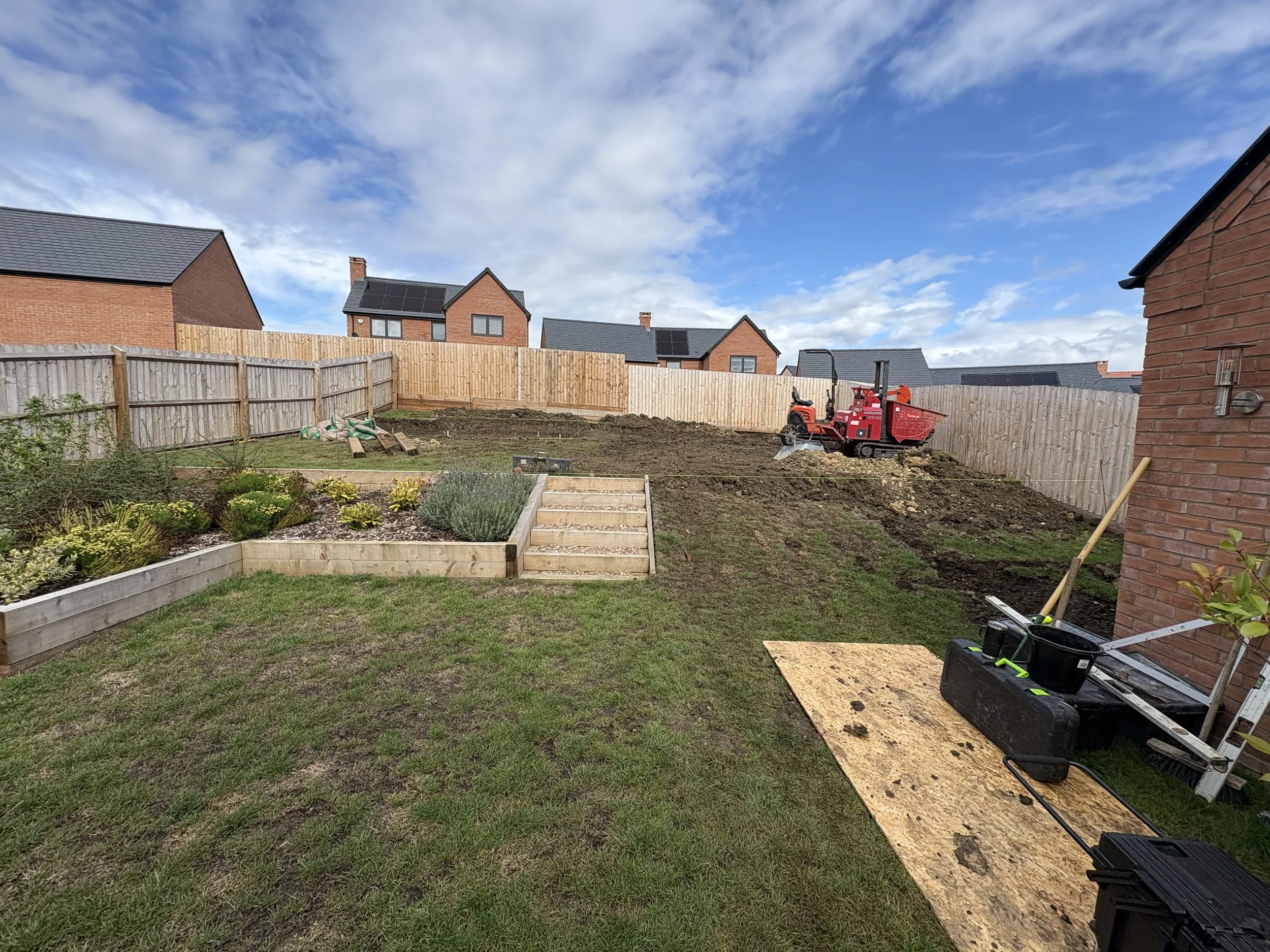 Backyard garden area under construction with raised garden beds, a small red construction vehicle, and building materials, with a wooden fence and houses under a partly cloudy sky.