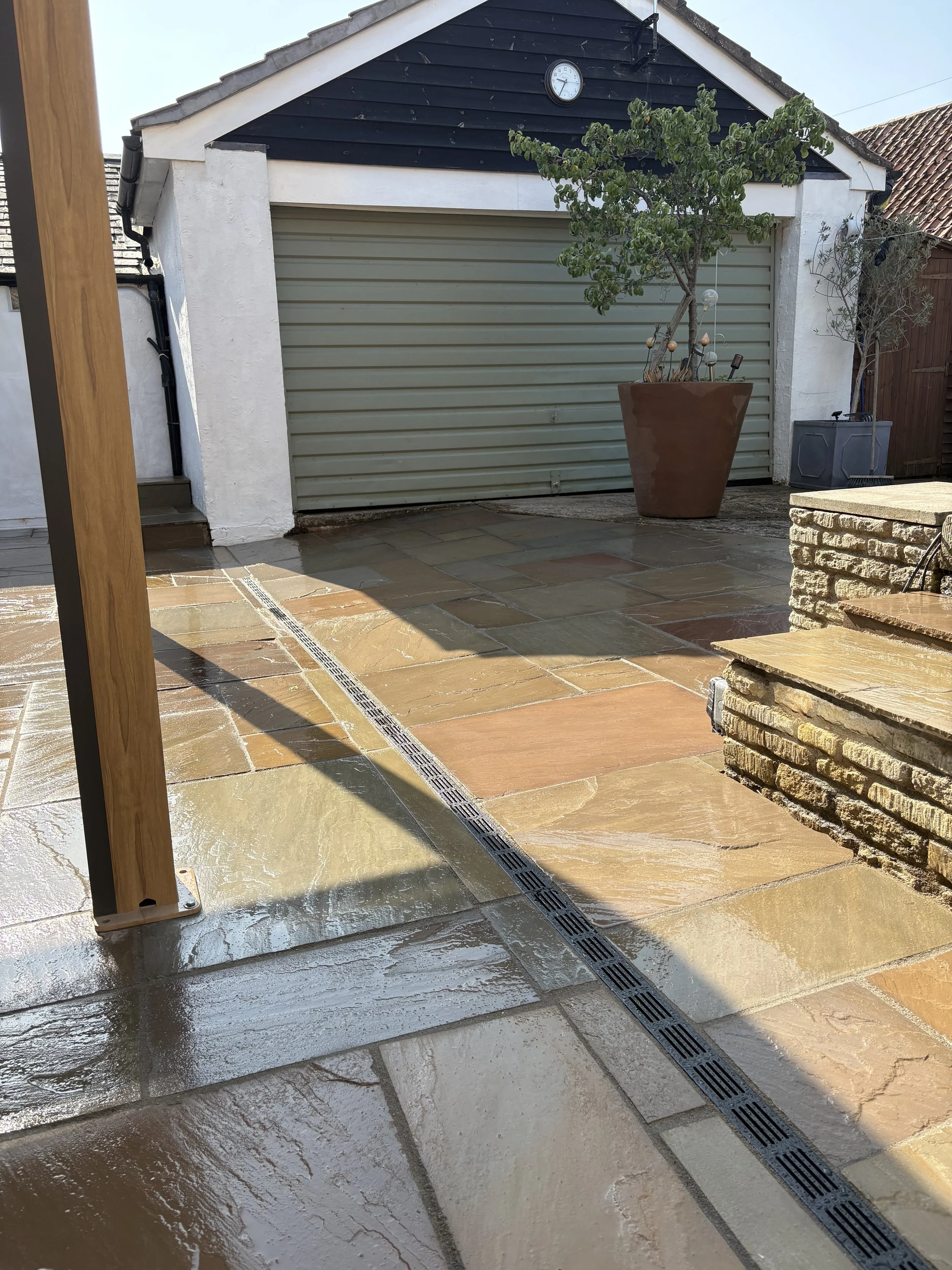An outdoor patio with wet stone tiles, a drain running through, a wooden post on the left, and a black garage door with a clock above it in the background. There are potted trees and a brick raised area on the right.