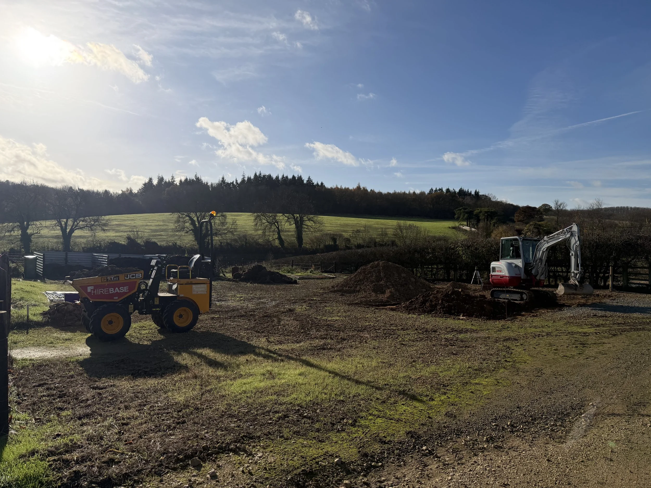 Construction site with a small excavator and a compact loader on a grassy field, with trees and a blue sky in the background.
