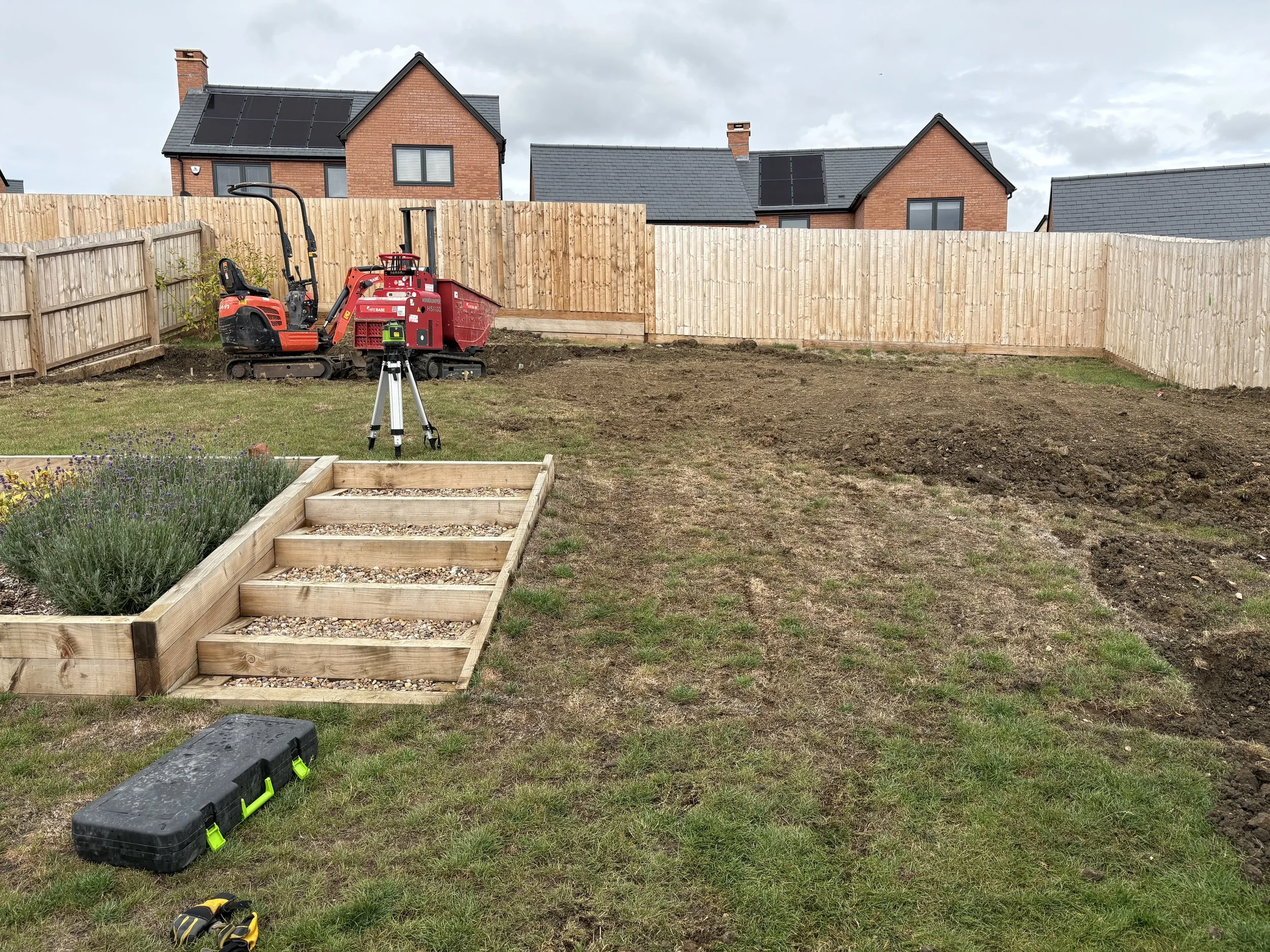 A backyard garden in the process of being landscaped, with a wooden raised flower bed, a small tracked excavator, a tripod with a level, and tools on the grass, surrounded by a wooden fence and neighboring houses with solar panels.