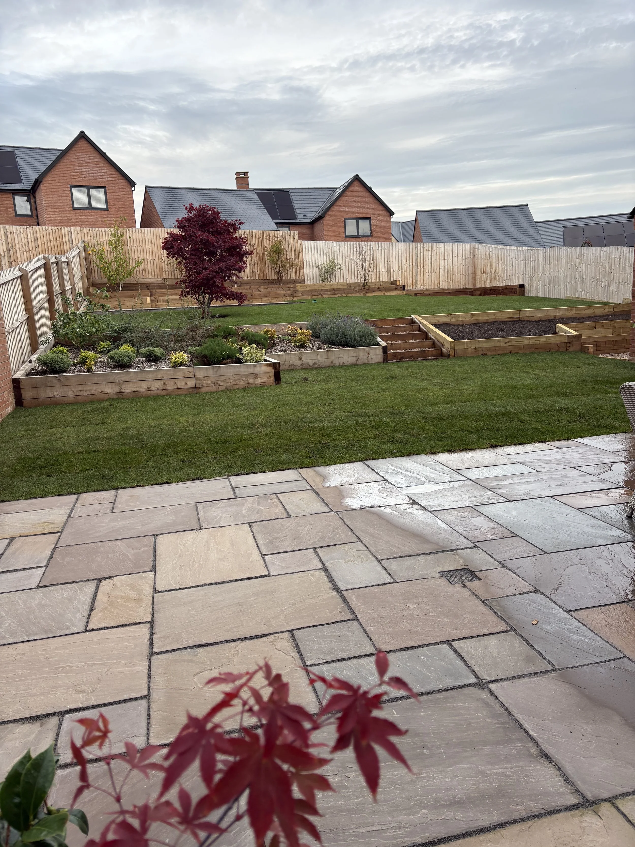 A backyard garden with a stone patio in the foreground, a grassy lawn, and multiple garden beds with plants and a small red leaves shrub, enclosed by a wooden fence. Houses with brick walls and gabled roofs are visible in the background under a partl