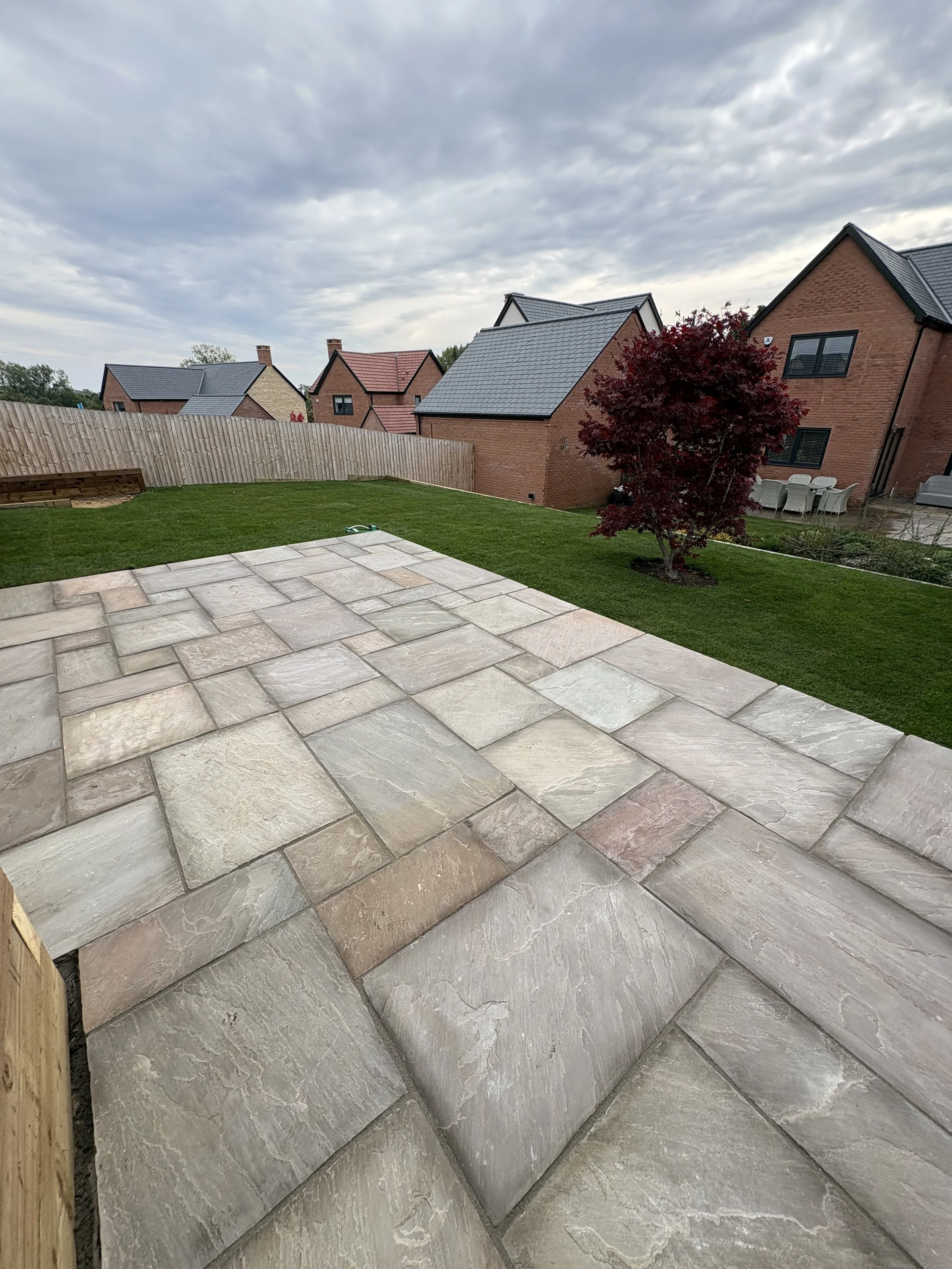 Backyard with a stone patio, green grass, a red tree, wooden fence, and neighboring houses under a cloudy sky.