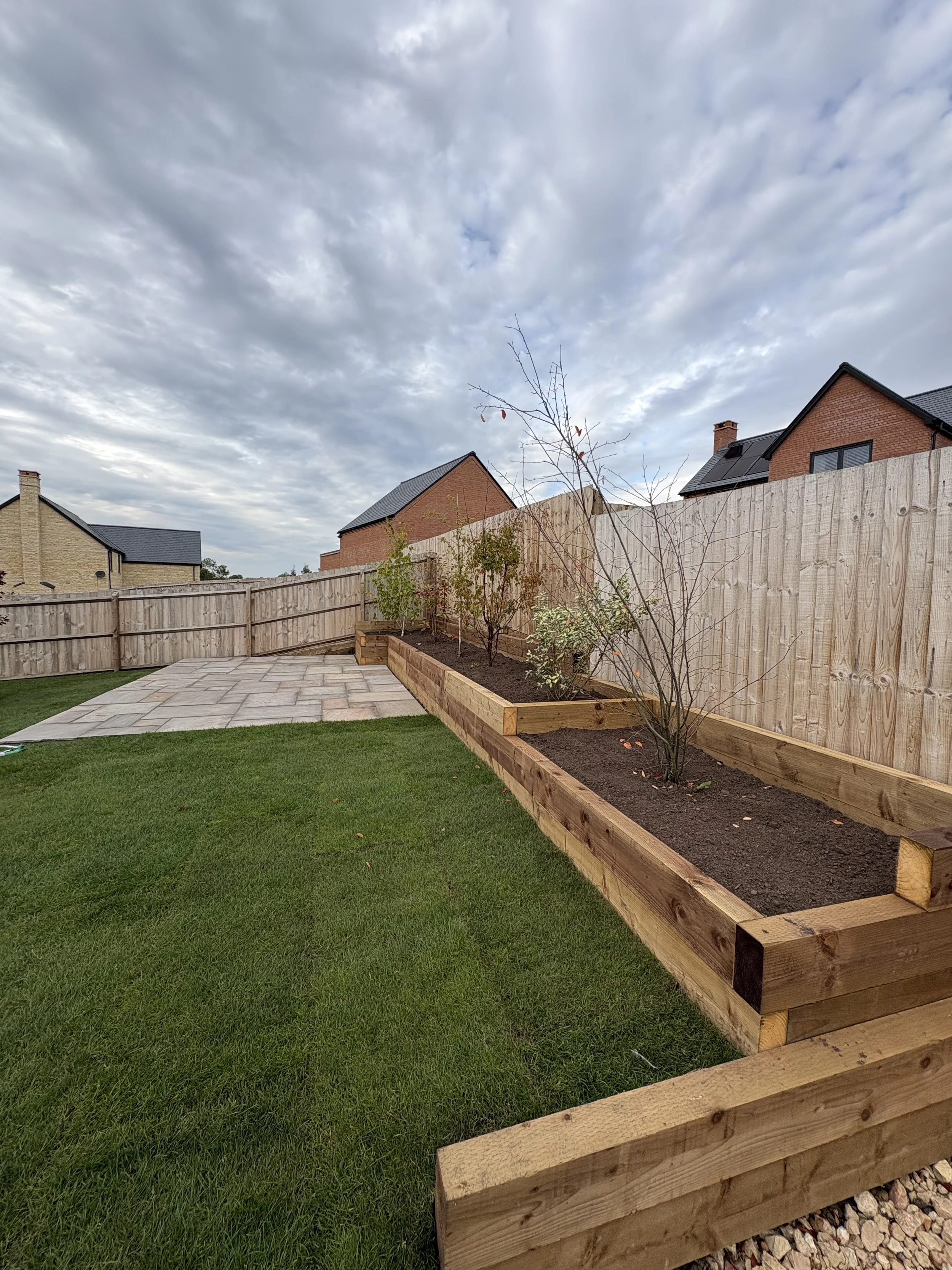 A backyard with a wooden raised garden bed containing plants, a paved patio, and a wooden fence under a cloudy sky.