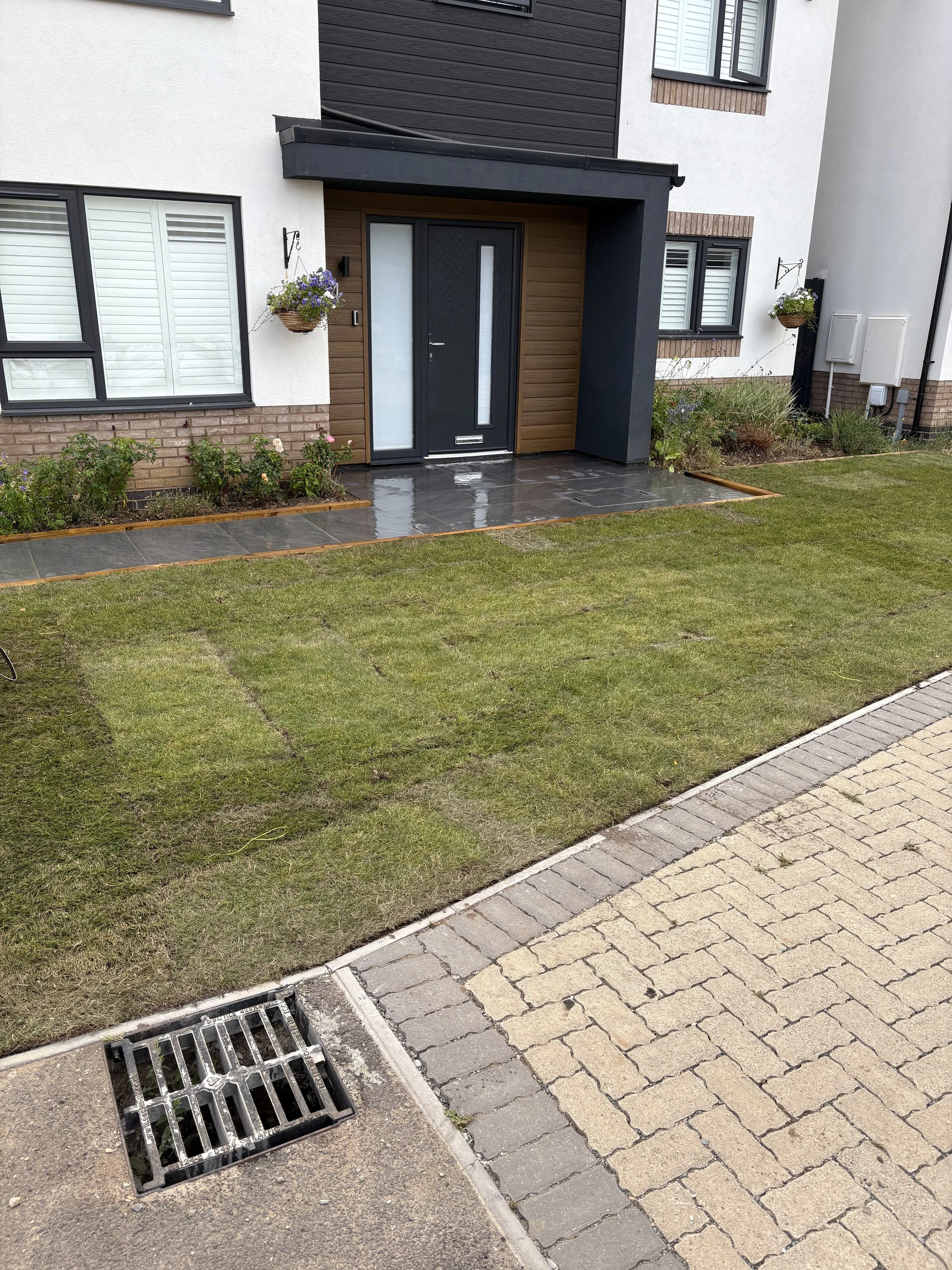 Front entrance of a modern house with a black door, grey tiled pathway, green lawn, flower bed, hanging flower baskets, brick-trimmed windows, and sidewalk with a drain grate.